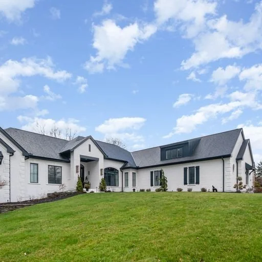 Modern white house with black roof and large windows, situated on a grassy lawn under a partly cloudy sky.