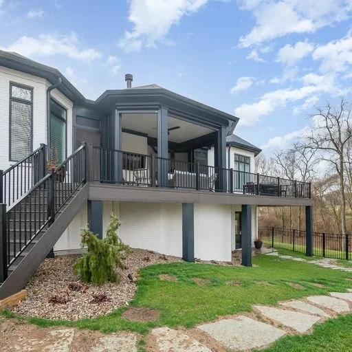 Exterior view of a two-story house with a balcony and stairs, surrounded by a lawn and a few trees.