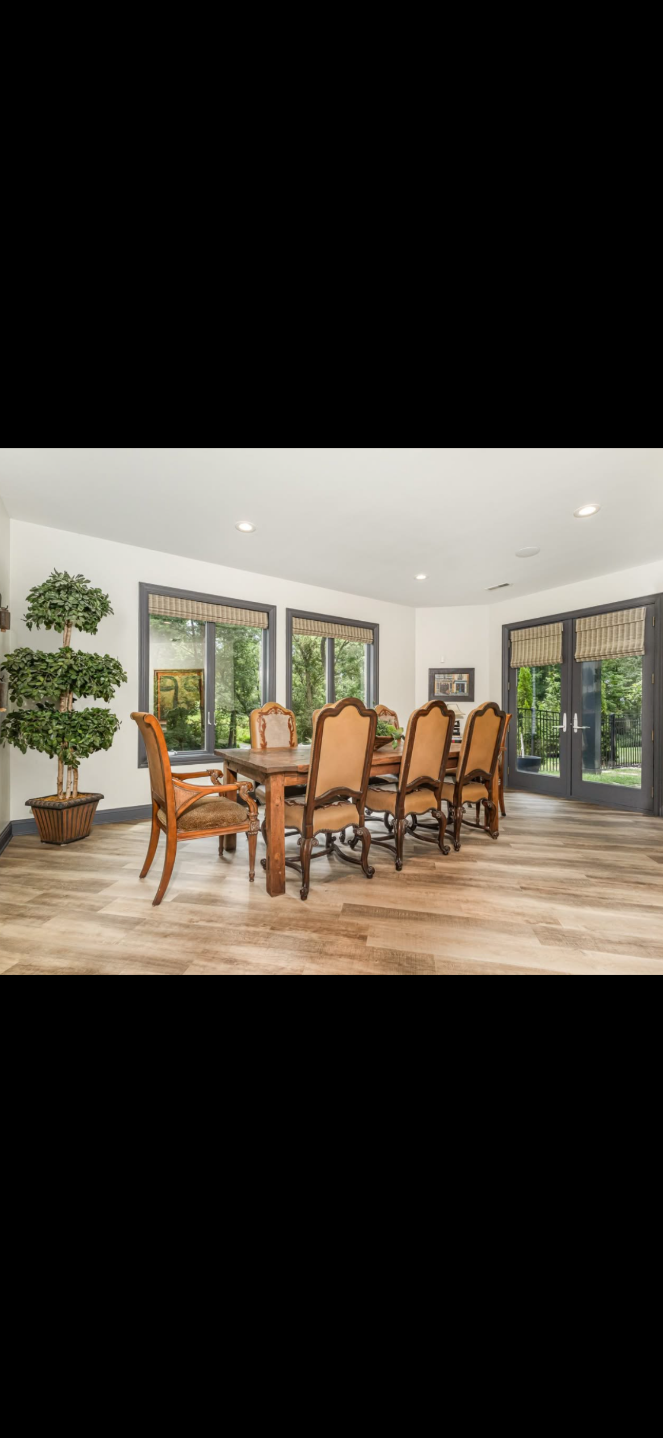 Dining room with wooden table, eight beige chairs with wooden frames, large windows, French doors, potted plant, hardwood flooring, framed artwork, and view of green outdoor landscape.