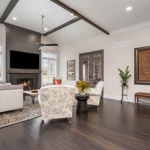 Living room with white walls, dark hardwood floors, and a modern fireplace with a television above. There are two patterned armchairs, a sofa, a black side table with a flower arrangement, a large window, and various wall art.