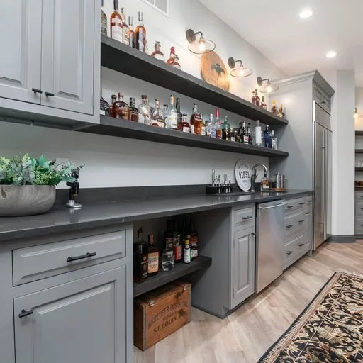 Gray kitchen with upper and lower cabinets, open shelves with liquor bottles, stainless steel dishwasher, countertop, and a decorative rug on wooden floor.