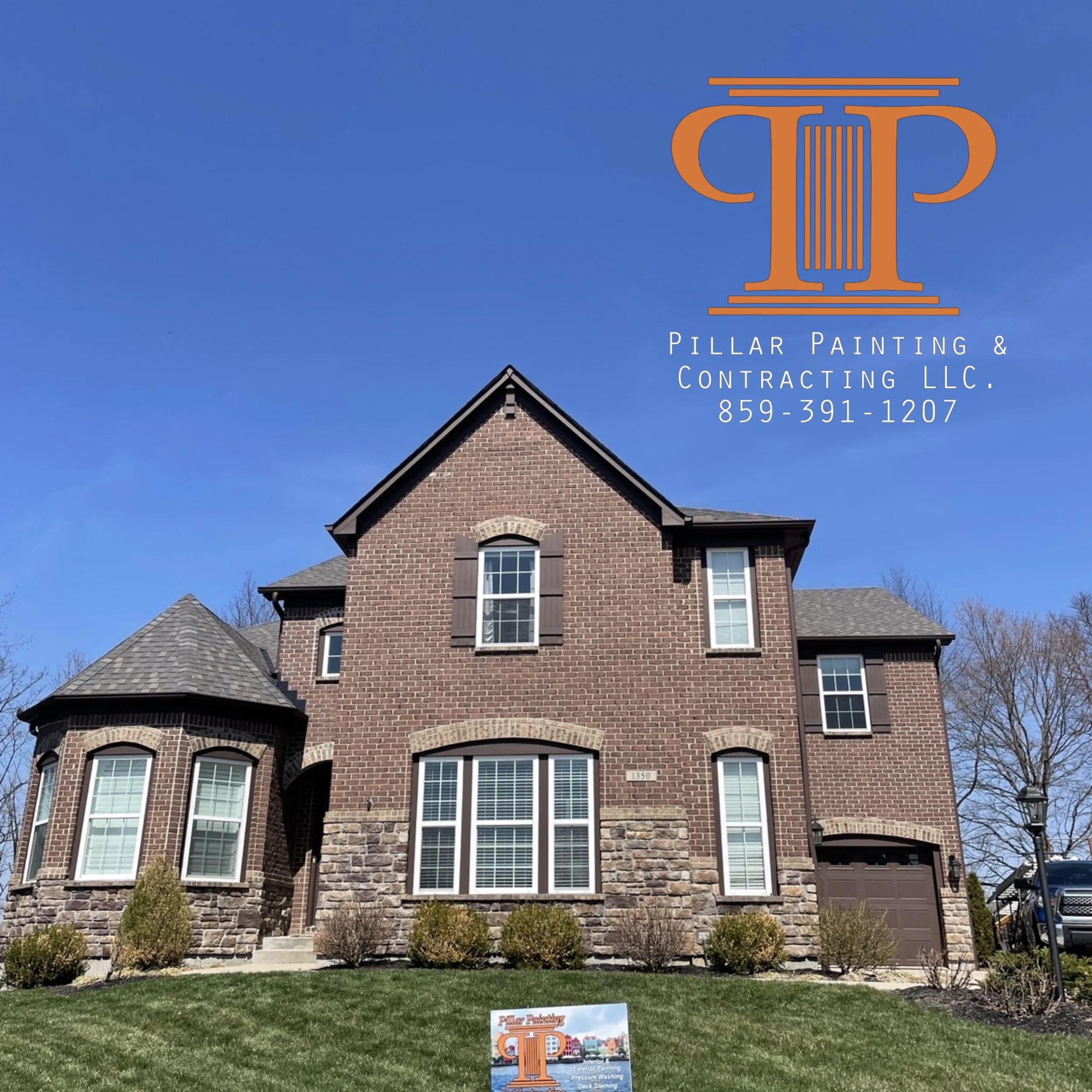 A large brick house with multiple windows and a steep roof, set against a clear blue sky. There is a sign in the front yard advertising pillar painting and contracting services.