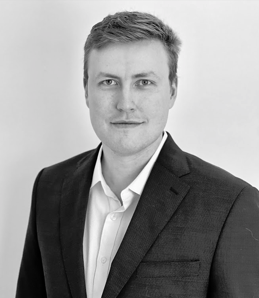 Black and white portrait of a young man wearing a suit and collared shirt, smiling slightly, against a plain background.