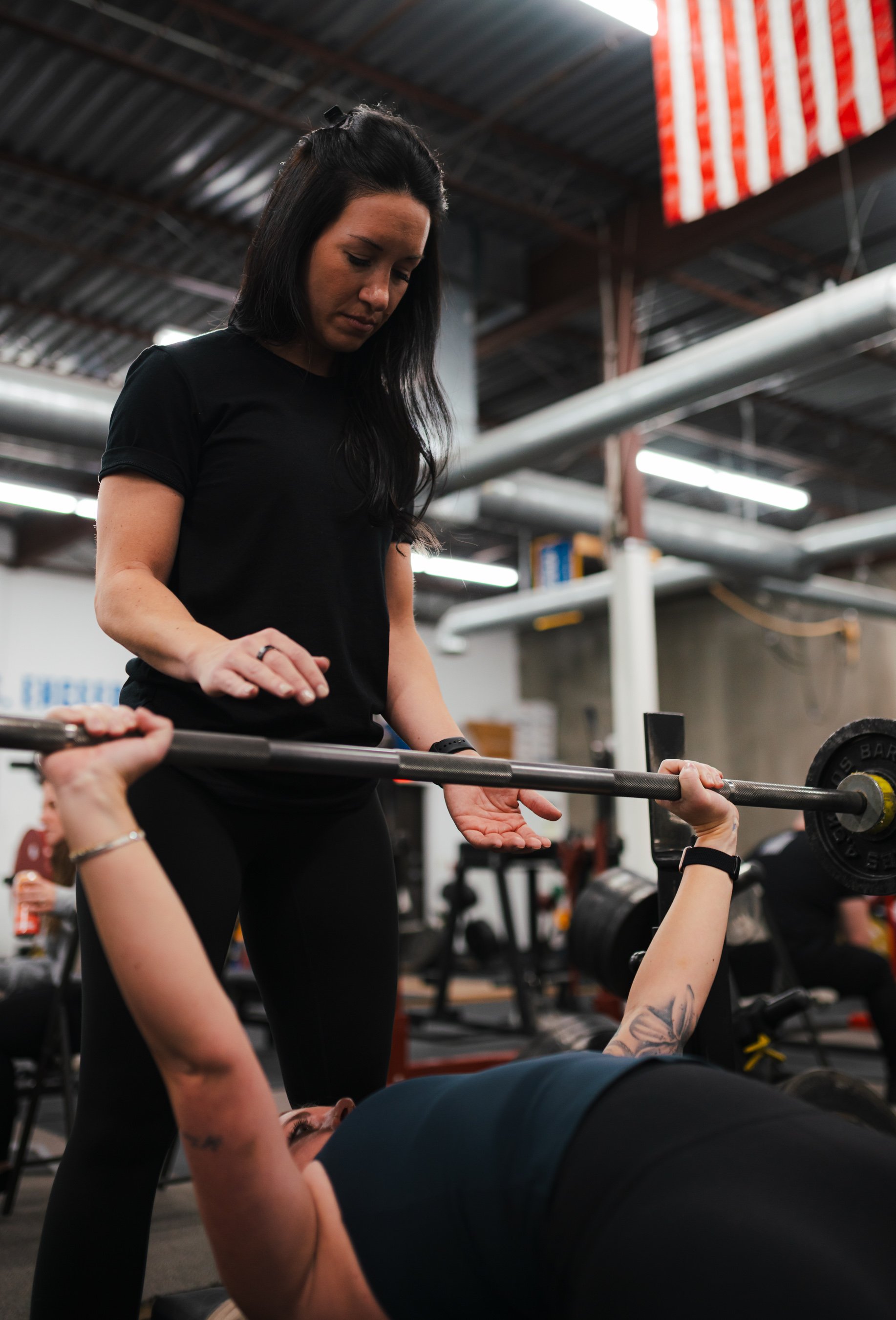 A woman spotting at a woman lying on a bench press in a gym with industrial ceiling and equipment.