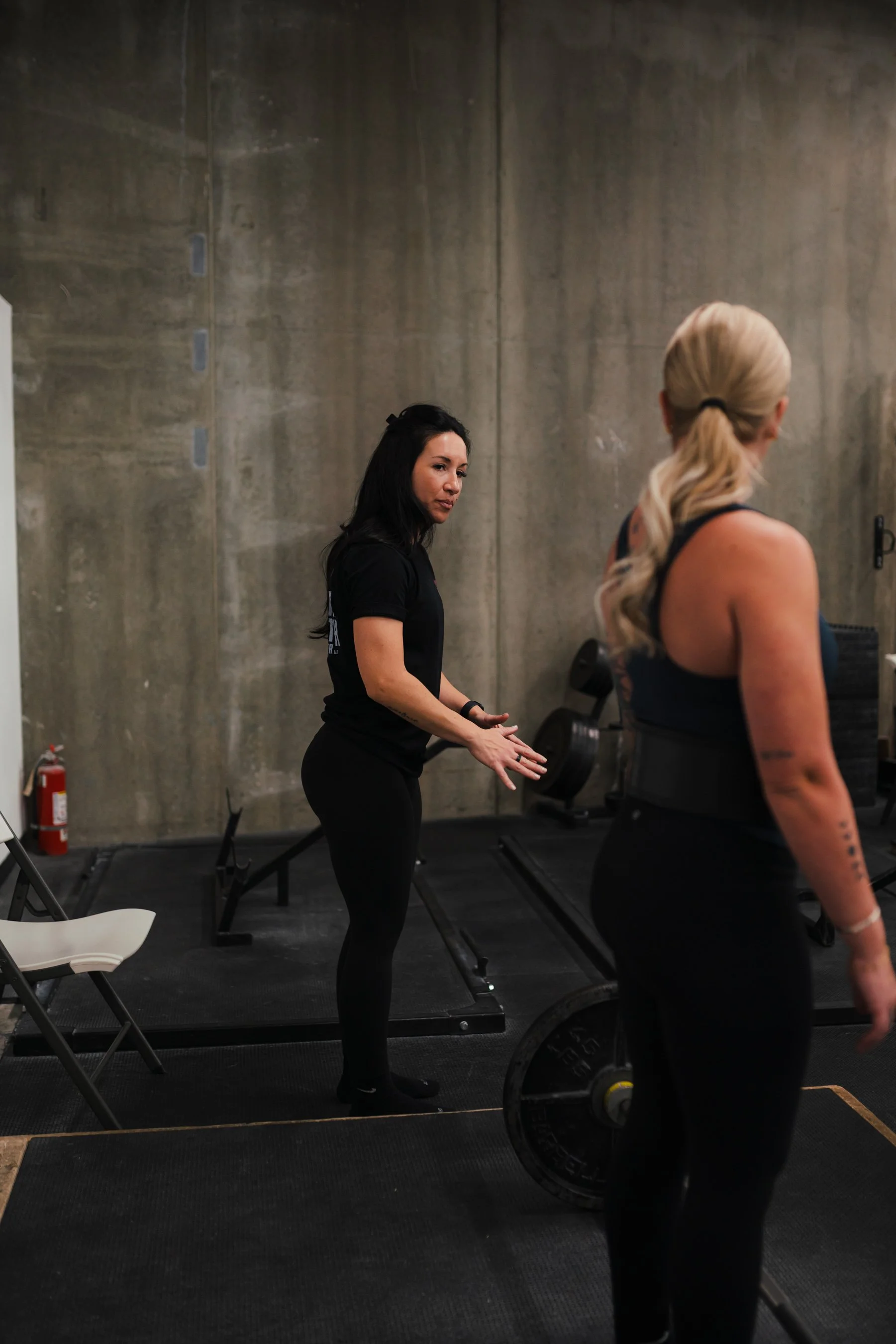 Two women in a gym with concrete walls, one with blonde hair in a ponytail facing away, the other with black hair and tattoos on her arm, standing near weights and gym equipment.