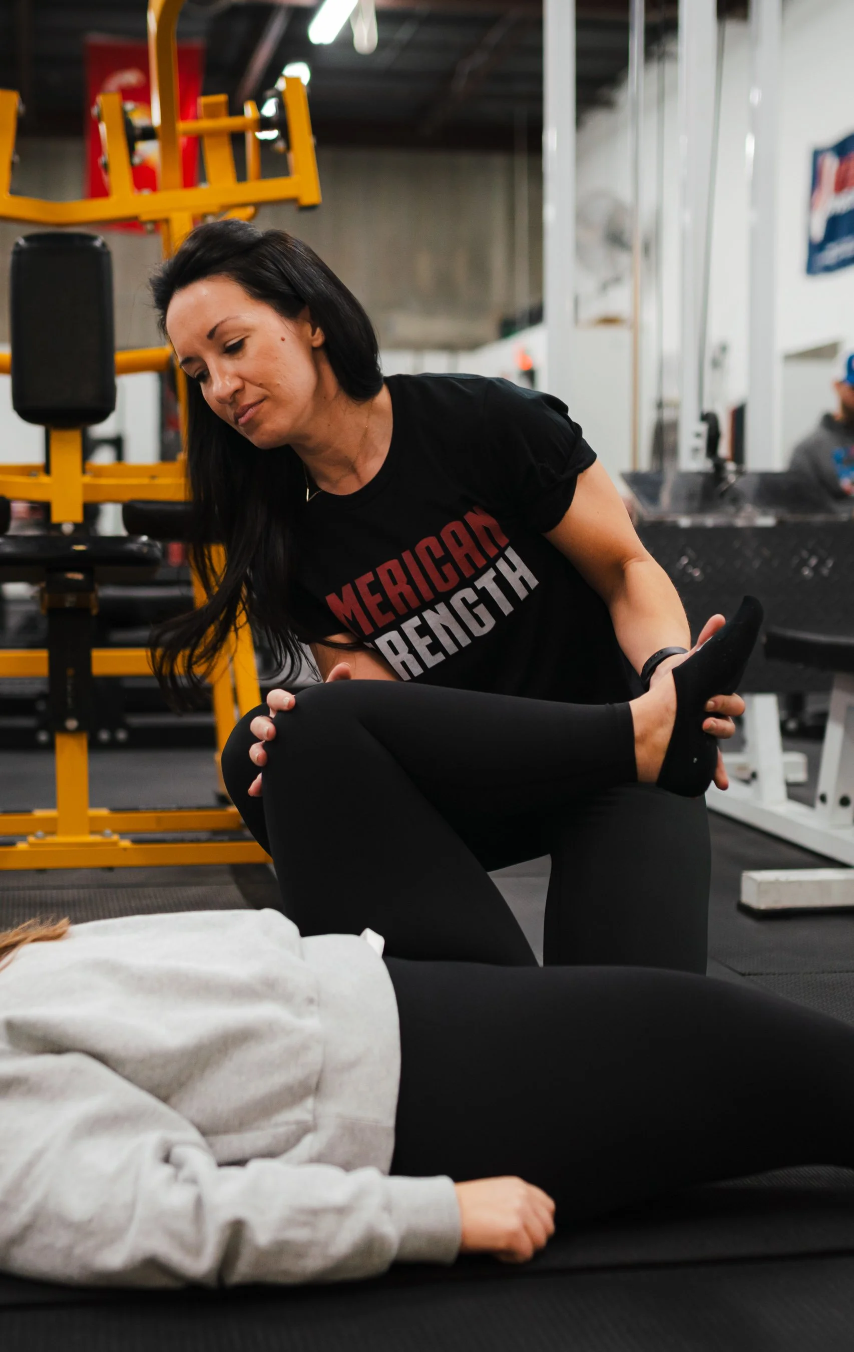 A woman in a black shirt performing a stretch exercise at a gym, with fitness equipment and a person lying down in foreground.