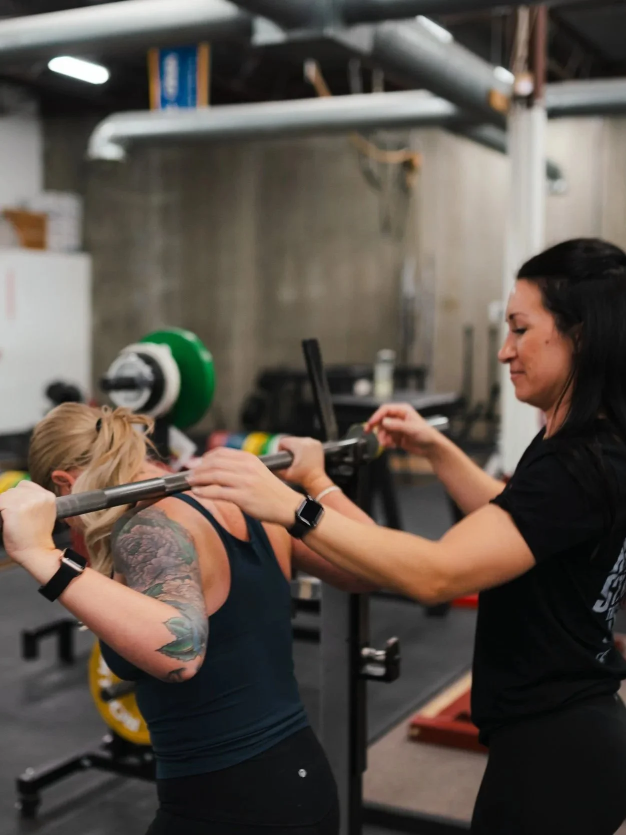 A woman with tattoos on her arm lifting weights with a barbell in a gym while another woman spots her.