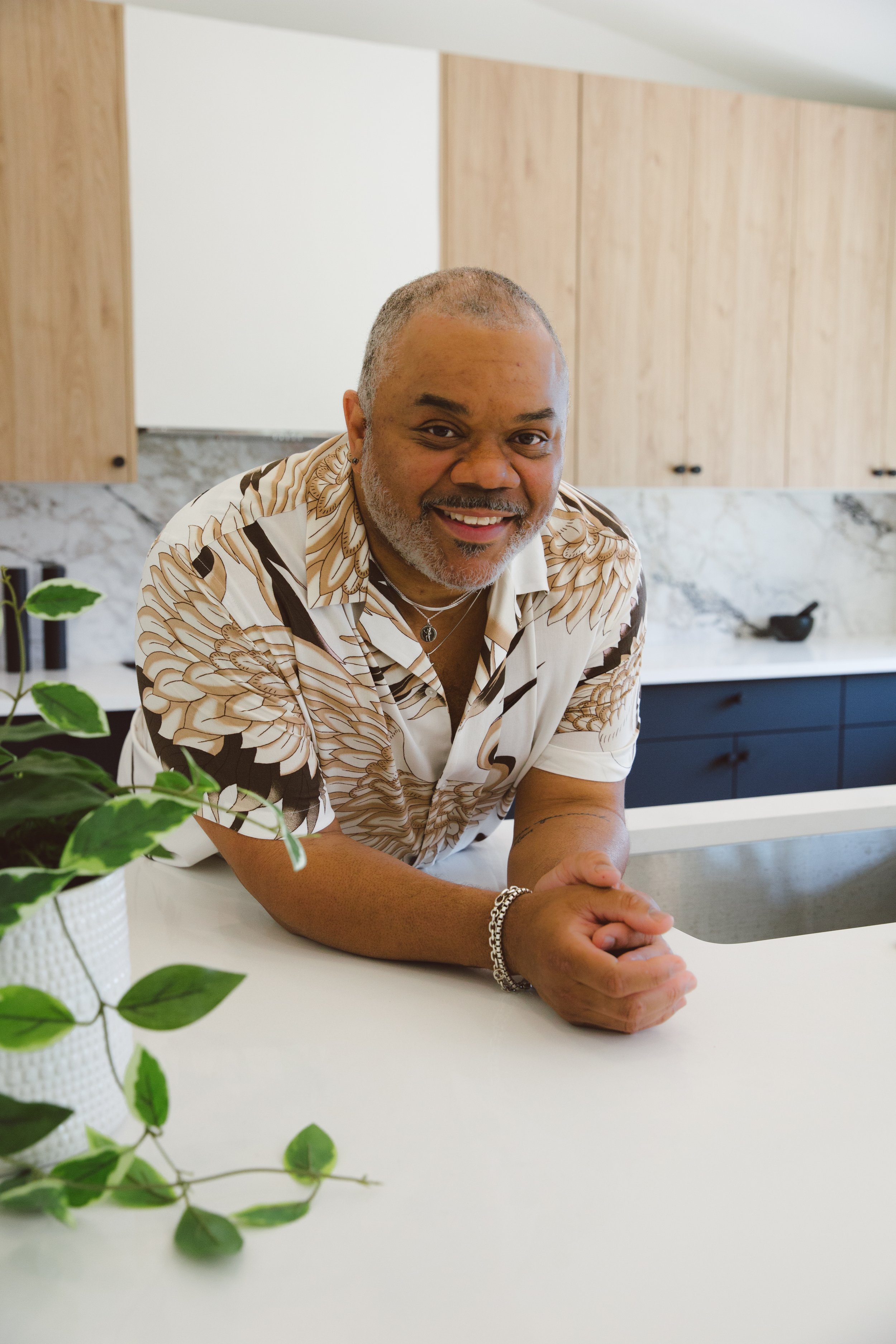 A smiling man with gray hair and beard, wearing a patterned shirt, leaning on a kitchen counter with greenery in a white pot in the foreground.
