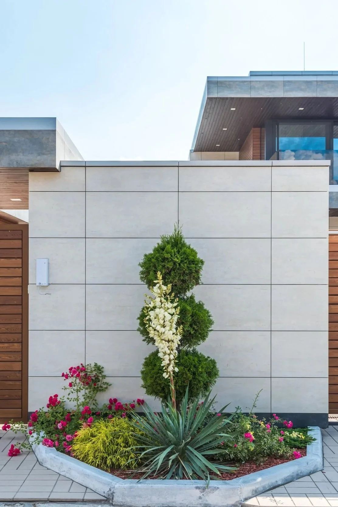 Front yard with a landscaped flower bed, small trees, and flowering plants in front of a modern house with beige, brown, and glass architectural elements.