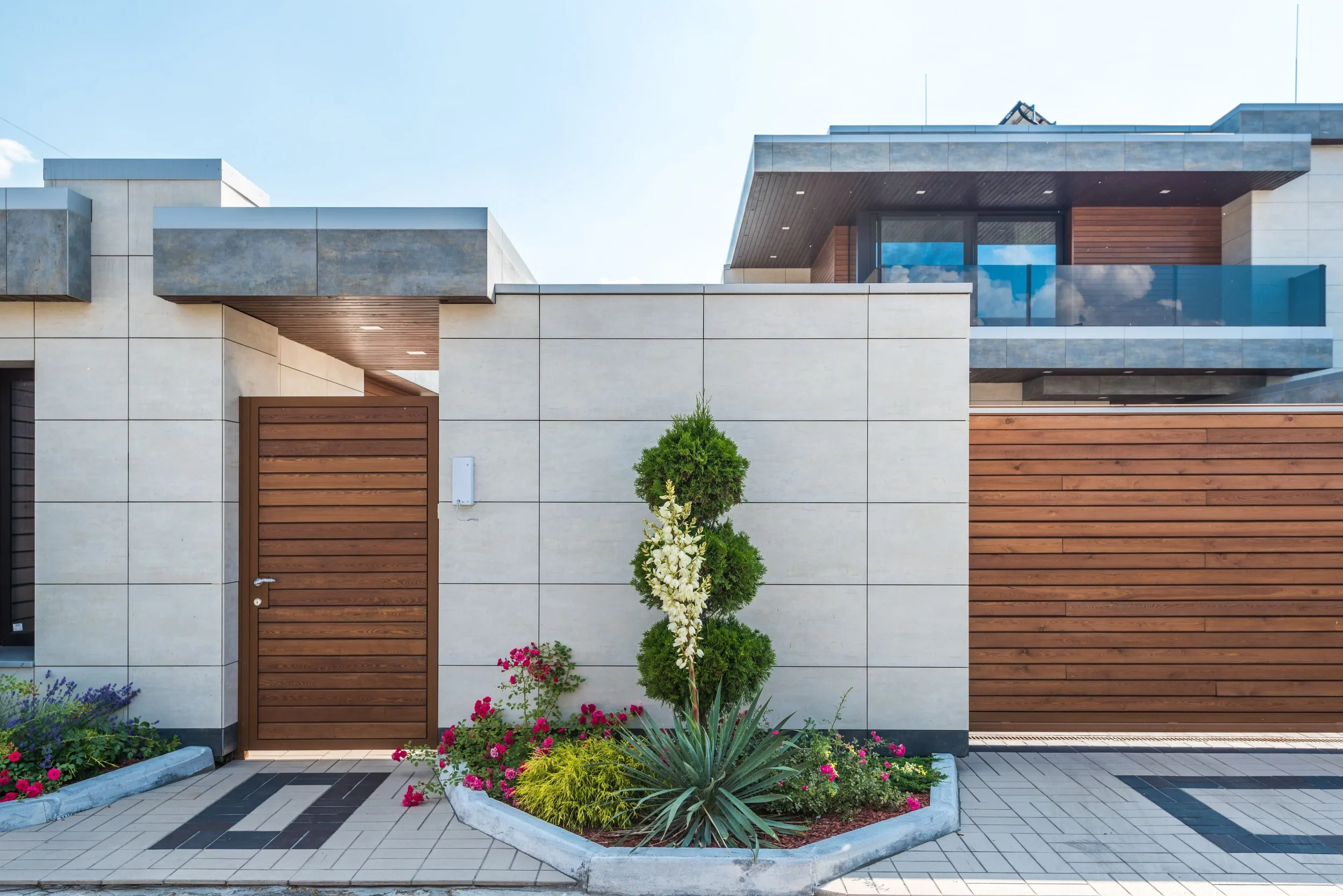 Modern house with a gate, a small landscaped garden with flowers, a tree, and a tiled walkway in front.