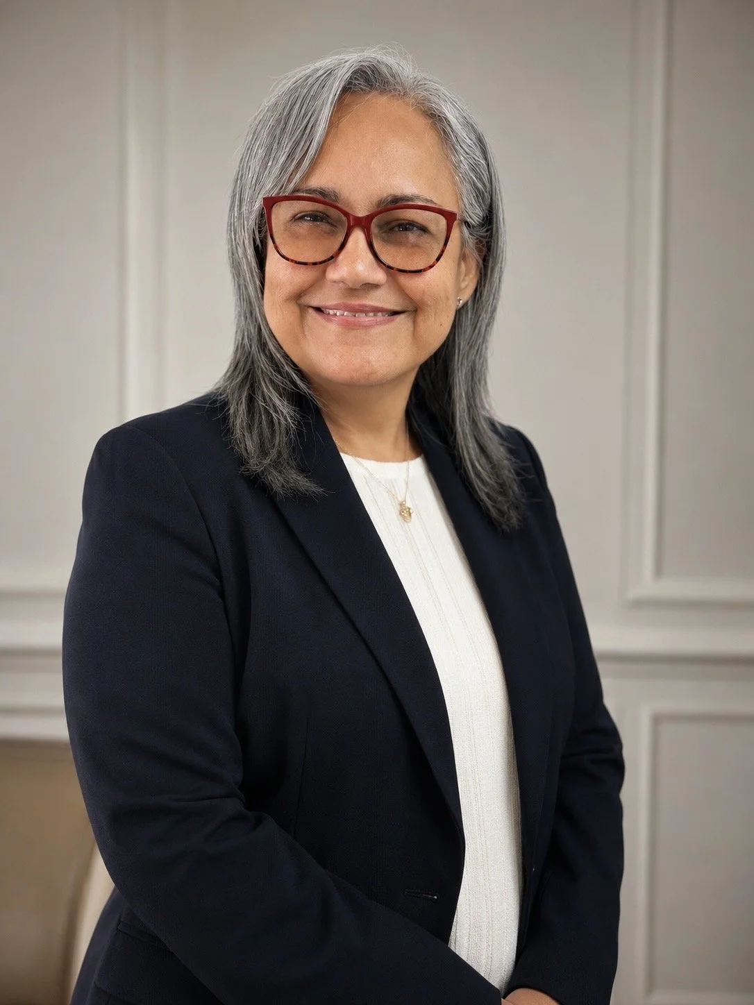A woman with gray and black curly hair, wearing red glasses, a white blazer with snowflake pattern, a coral top, and a gold necklace, standing against a plain light background. Miami Florida, telehealth, Spanish, mental health