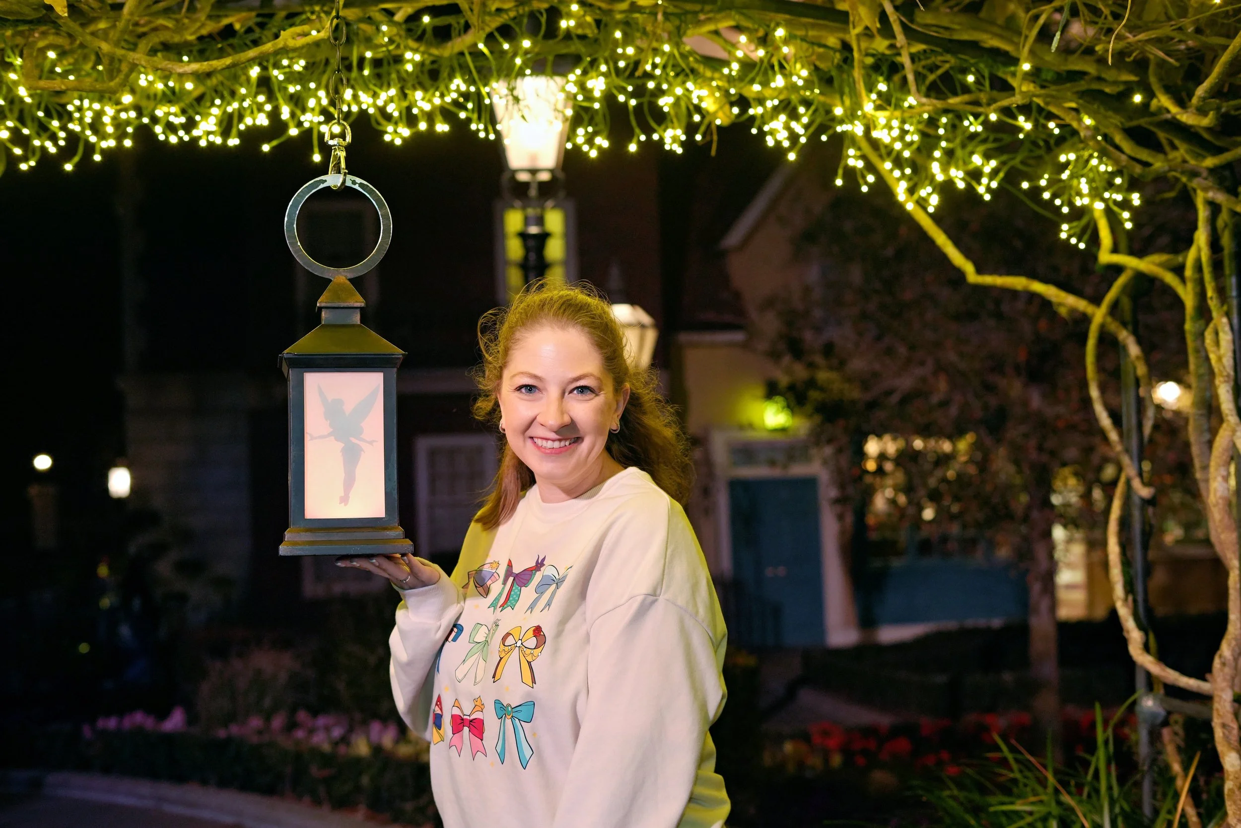 A woman smiling outdoors at night holds a lantern with a fairy silhouette inside, under string lights with a house and trees in the background.