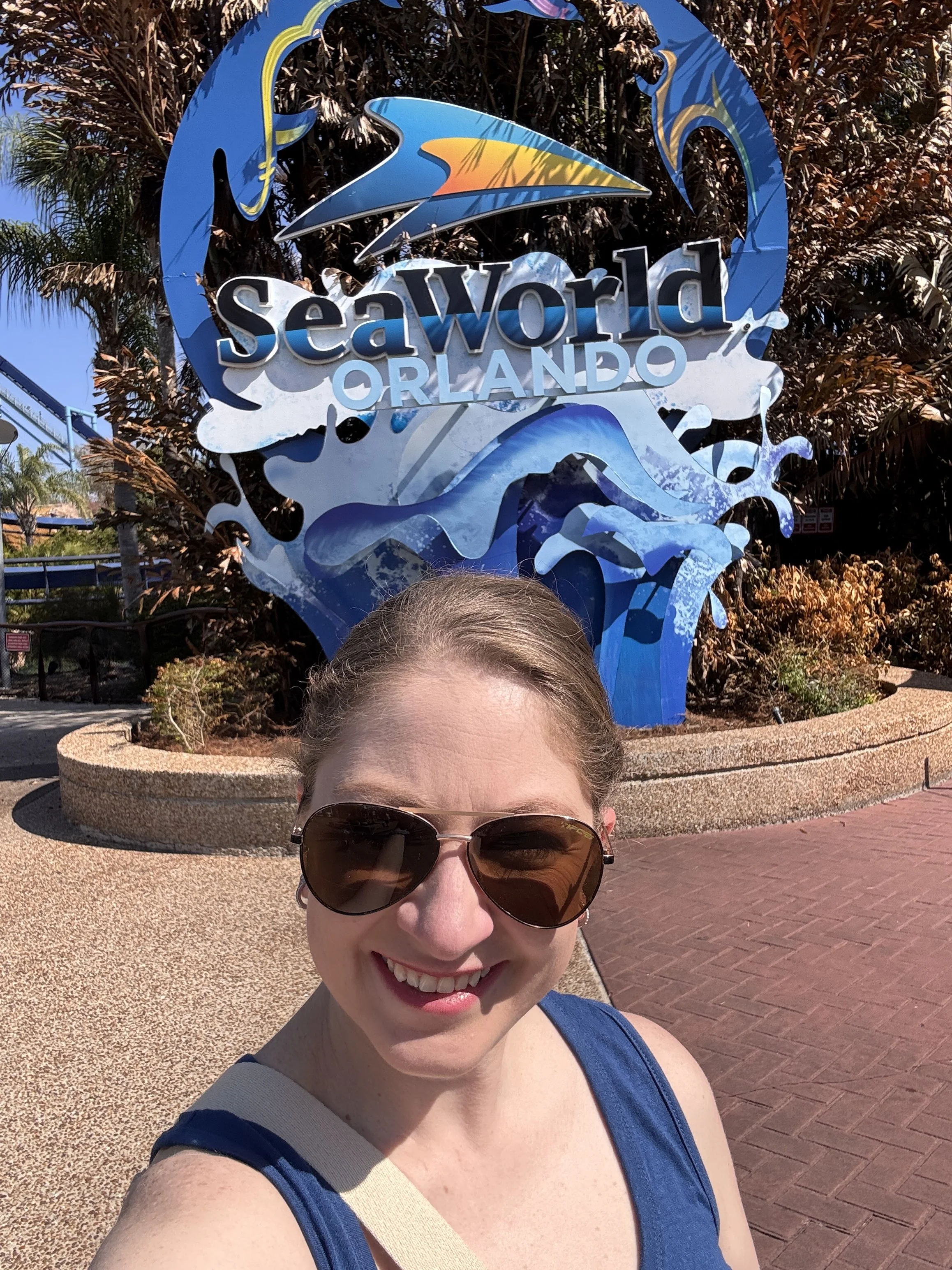 A smiling woman with sunglasses taking a selfie in front of the SeaWorld Orlando sign on a sunny day.