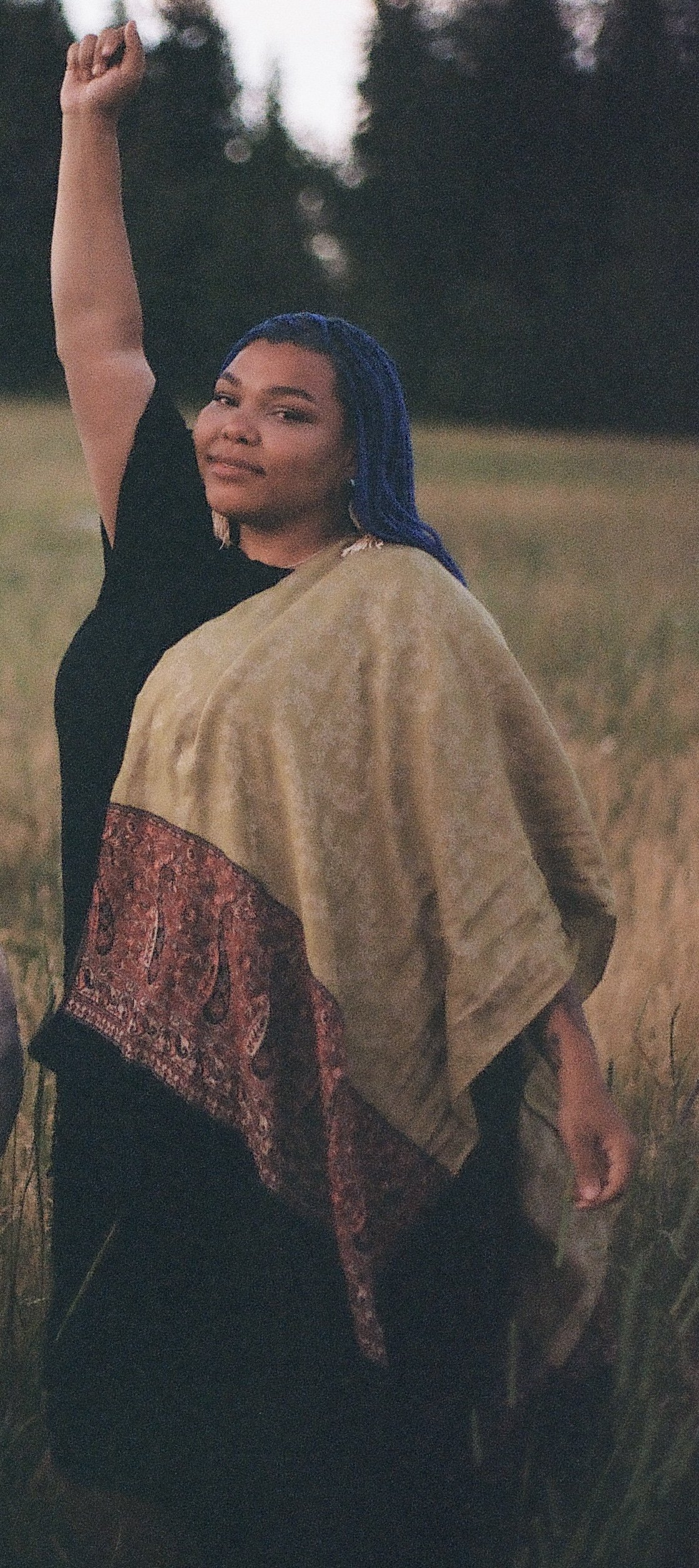 A woman with blue braided hair standing in a field at dusk, raising her right arm with a clenched fist, wearing a black top and a multicolored shawl.