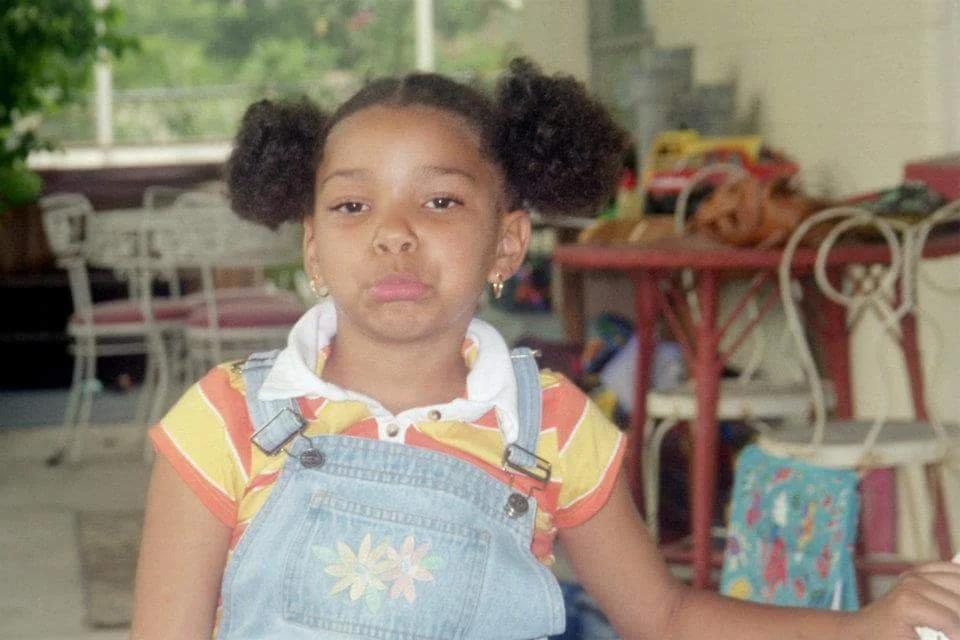 Young girl with puffy hair wearing a yellow and orange striped shirt and denim overalls, seated indoors.