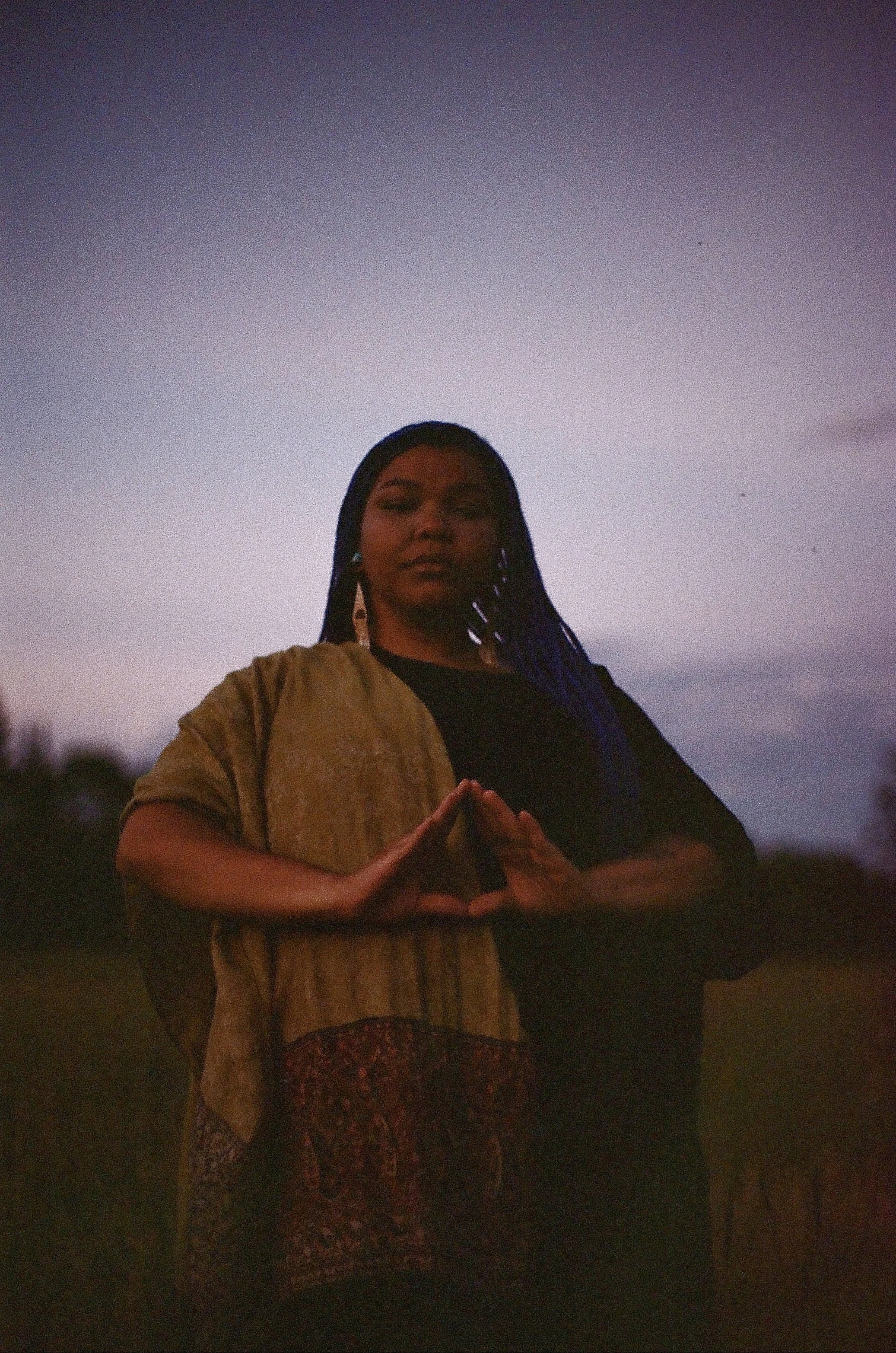 A woman standing outdoors during dusk, making a hand gesture that resembles a heart, with her fingers united at the fingertips, and wearing a beige jacket with a patterned dress underneath. She has long braided hair and a calm facial expression.