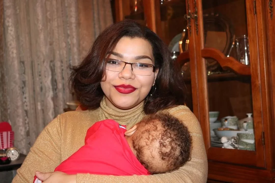 A woman wearing glasses and red lipstick is holding a baby wrapped in a red blanket. They are in a room with wooden cabinets and dishware in the background.
