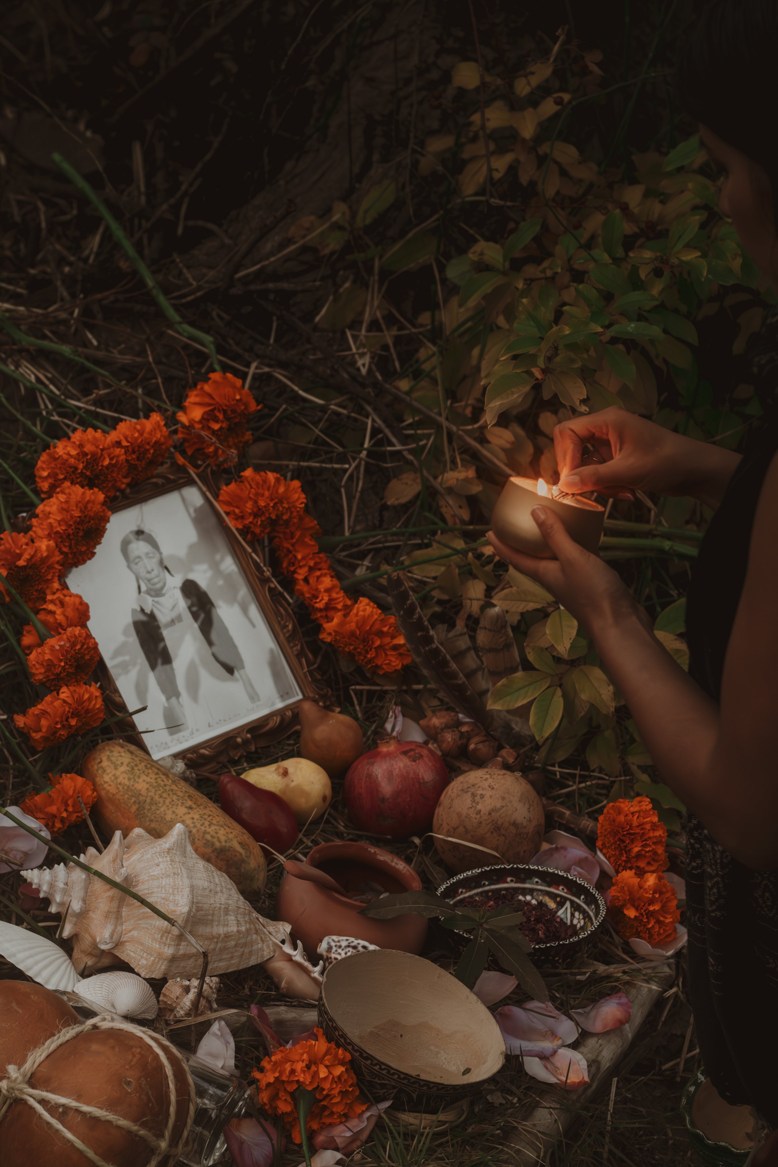 A person lighting a candle at a grave decorated with orange marigold flowers, with objects like seashells, fruits, and a framed black-and-white photograph nearby.