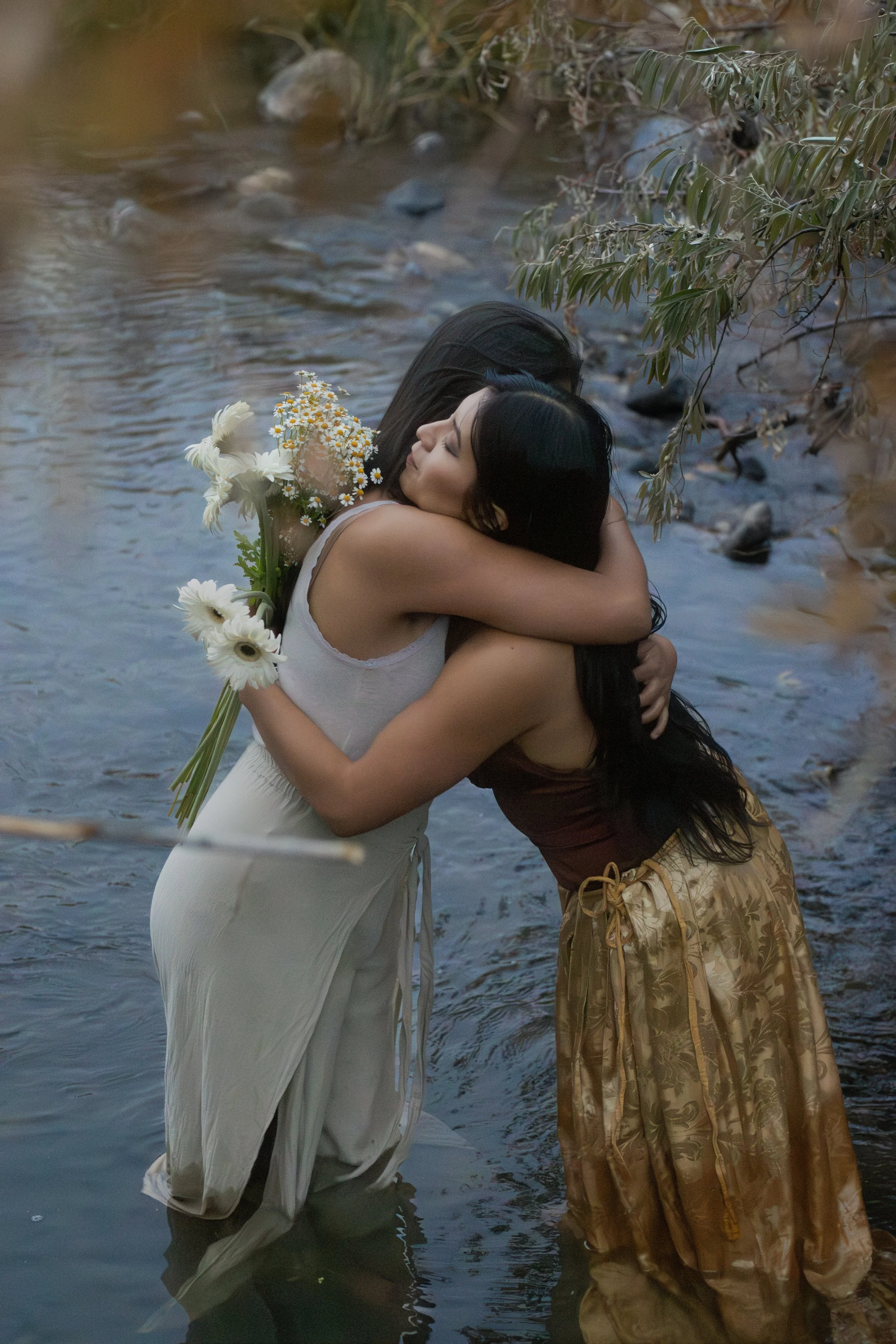Two women embrace in a shallow river, one holding a bouquet of white daisies. The woman in a white dress has her eyes closed, and the other woman, in a patterned skirt, cradles her. Trees line the riverbank.
