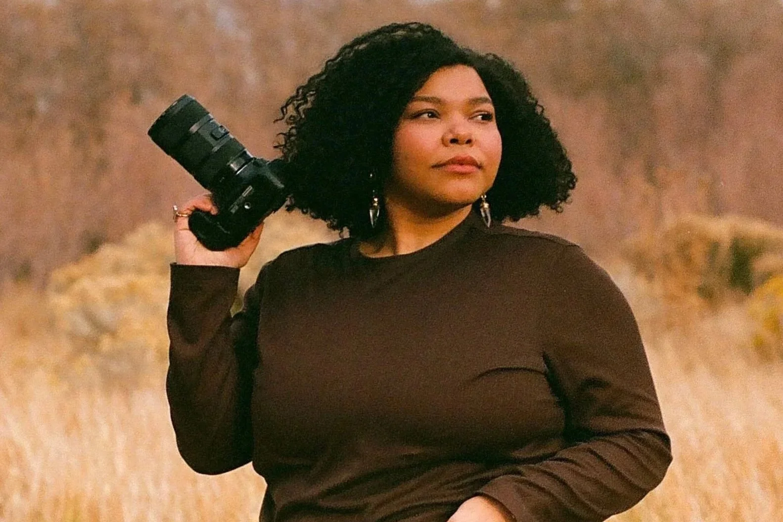A woman with curly black hair holding a camera on her shoulder, standing outdoors with a blurred natural background.