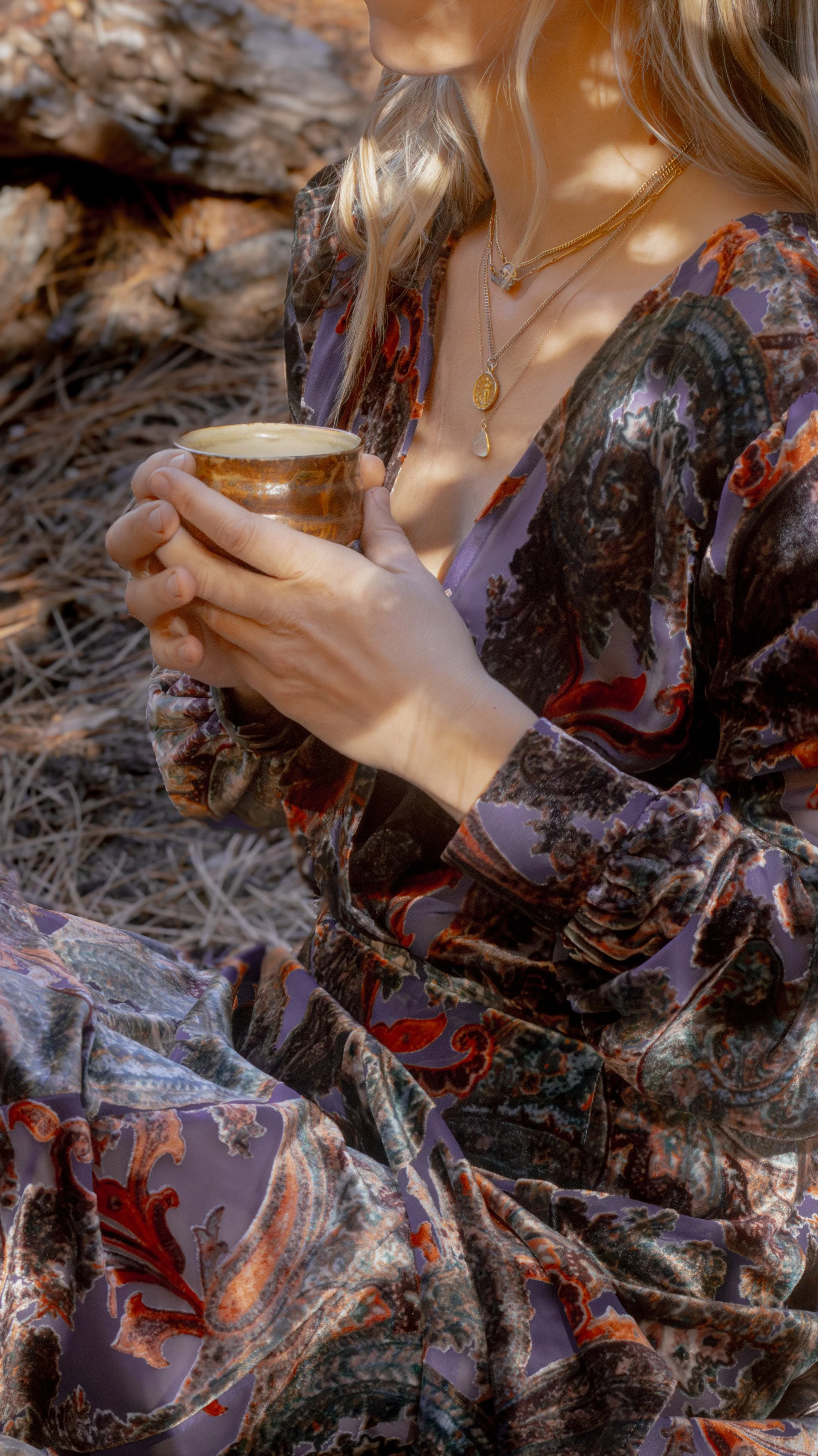 A woman wearing a colorful, patterned dress holding a ceramic cup with both hands, sitting outdoors on a bed of dry leaves and pine needles.