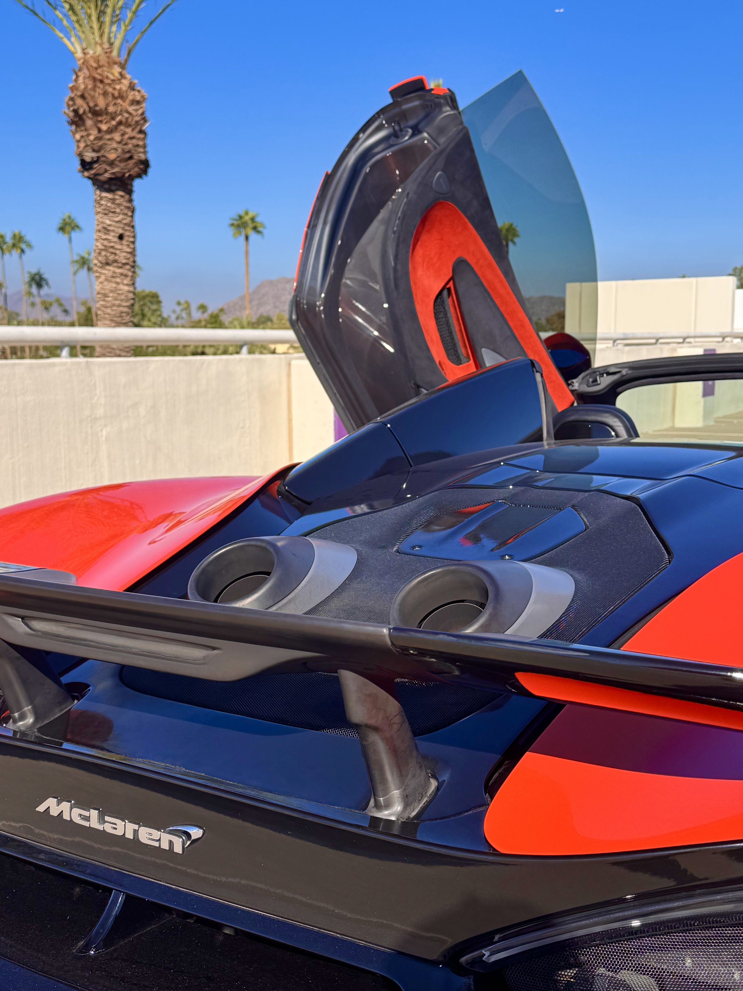 Close-up of a tuned and straight-piped Helios orange McLaren 600LT spider exotic sports car with its butterfly doors open, revealing dual top-mounted exhaust ports, parked outdoors under a blue sky with palm trees in the background.