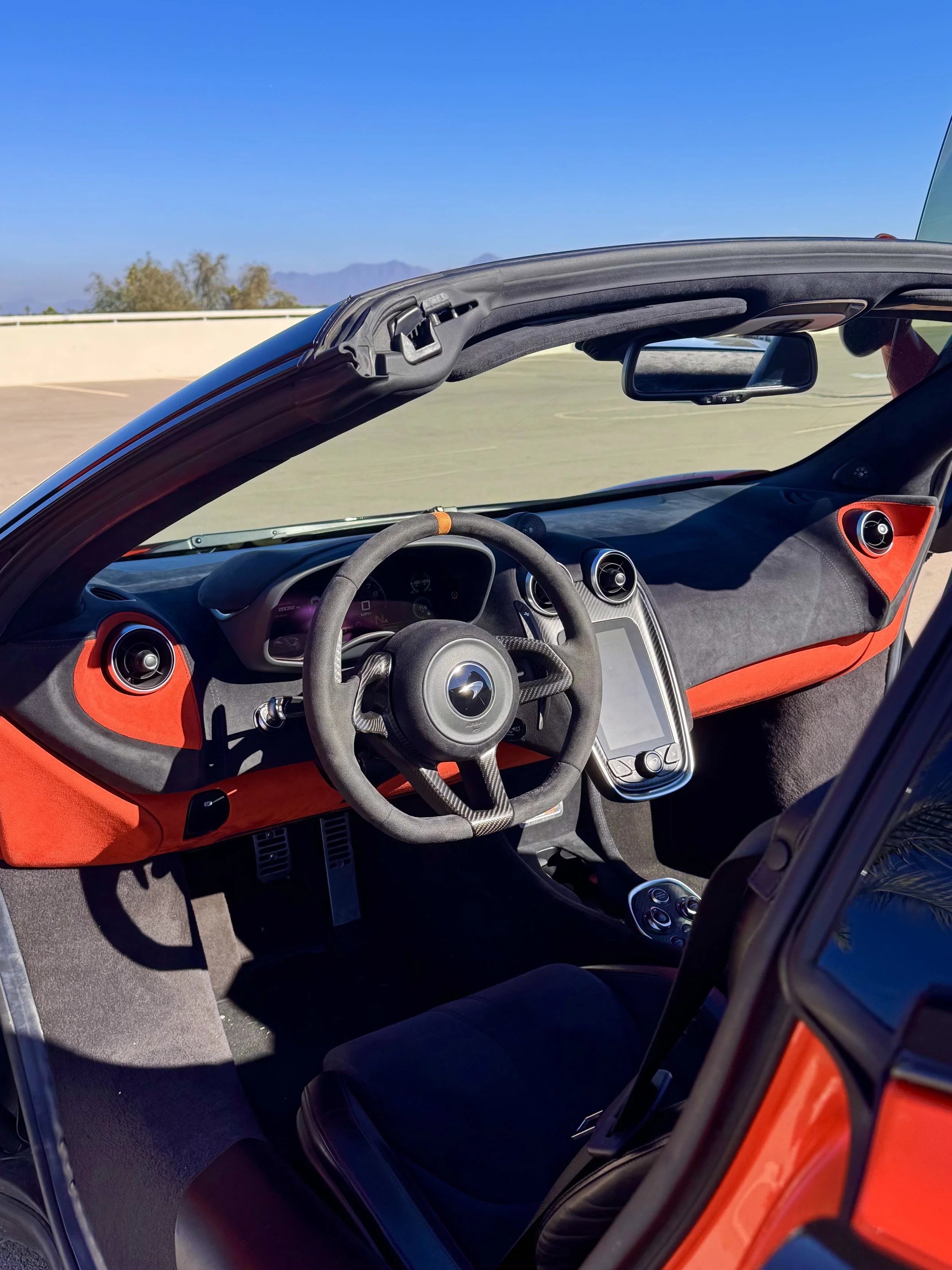 Interior view of a tuned and straight-piped Helios orange McLaren 600LT spider exotic sports car with a racing steering wheel, digital display, and touchscreen interface, parked in an open area under a clear blue sky.