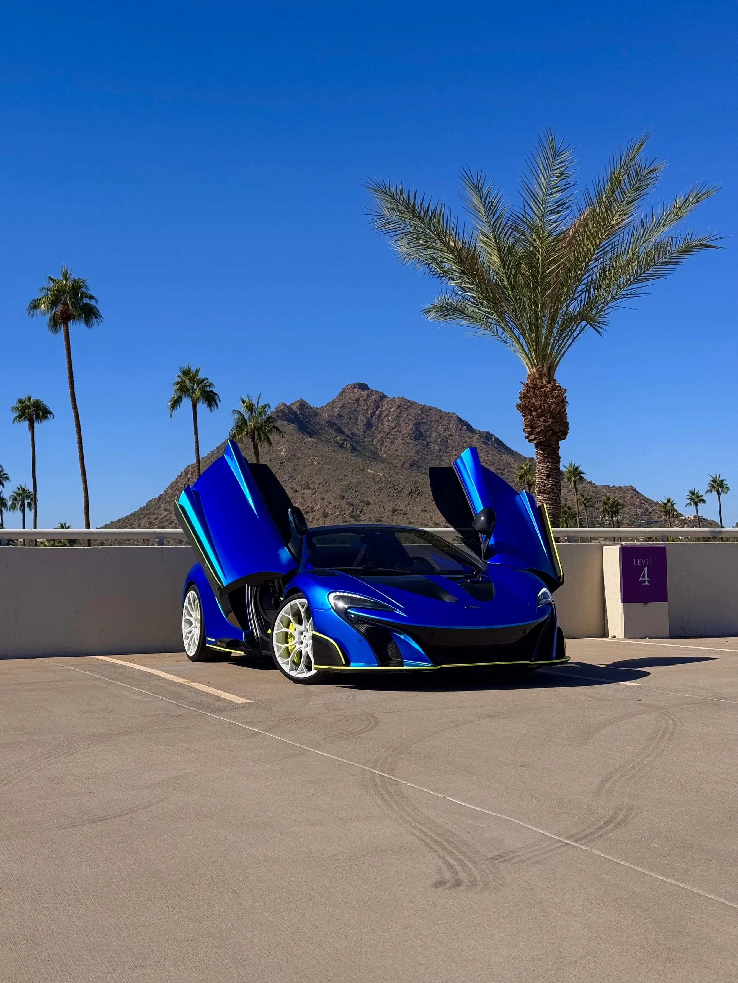 A blue McLaren 650S Spider with butterfly doors open parked on a concrete level in a desert-like area with palm trees and mountainous terrain in the background.