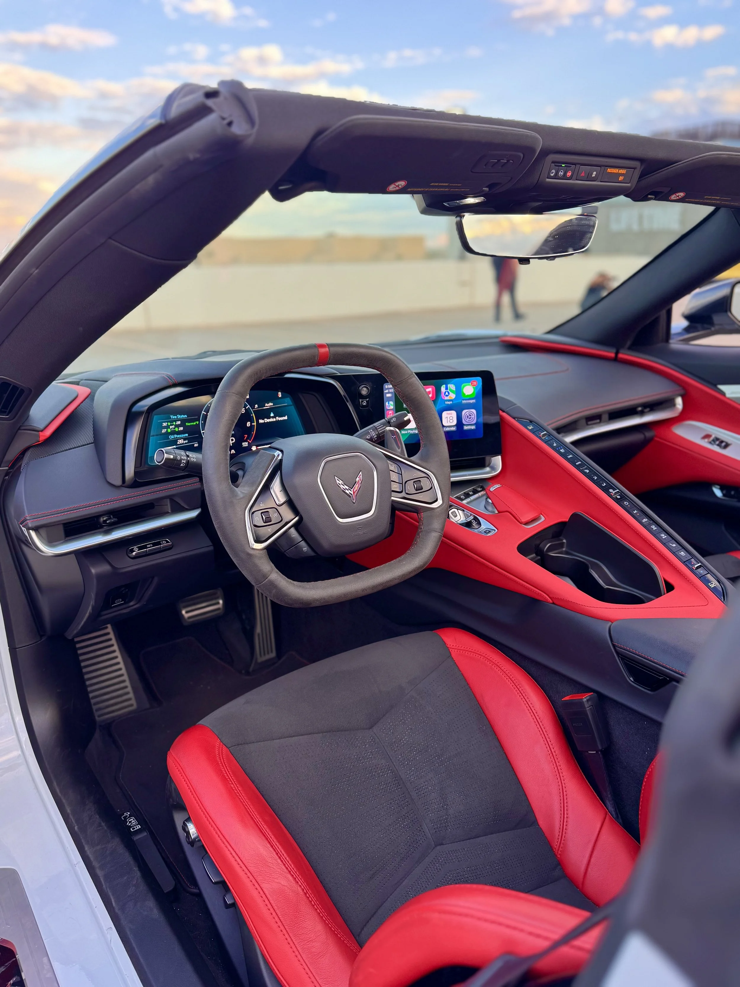 Interior of a White Chevrolet Corvette C8 convertible sports car with a black and red color scheme, featuring a steering wheel, digital dashboard, and touchscreen display, with an outdoor background visible through the windshield.