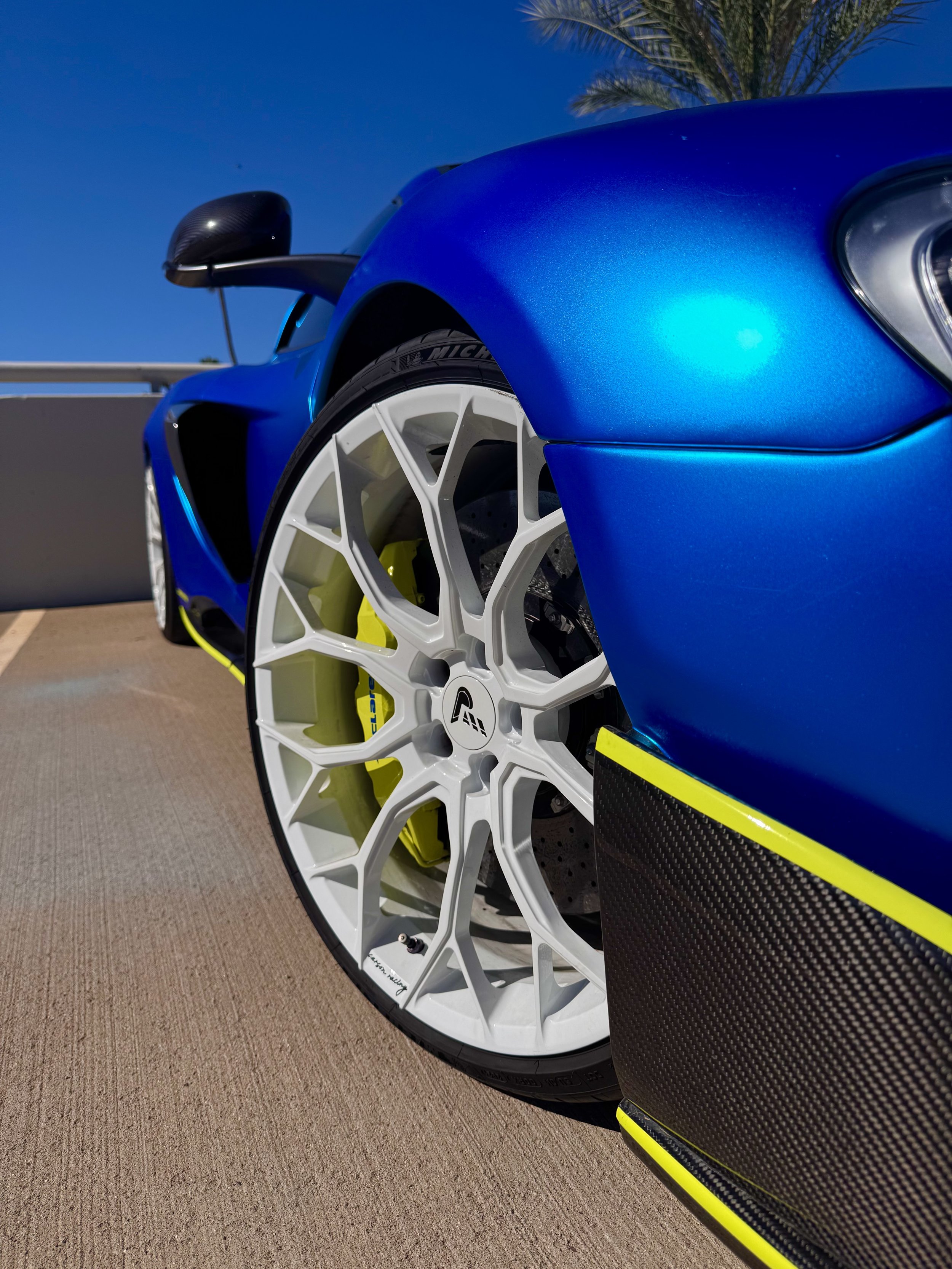 Close-up of a blue McLaren 650S Spider's front wheel and tire, including a white alloy wheel, yellow brake caliper, and carbon fiber side panel, with a palm tree and blue sky in the background.