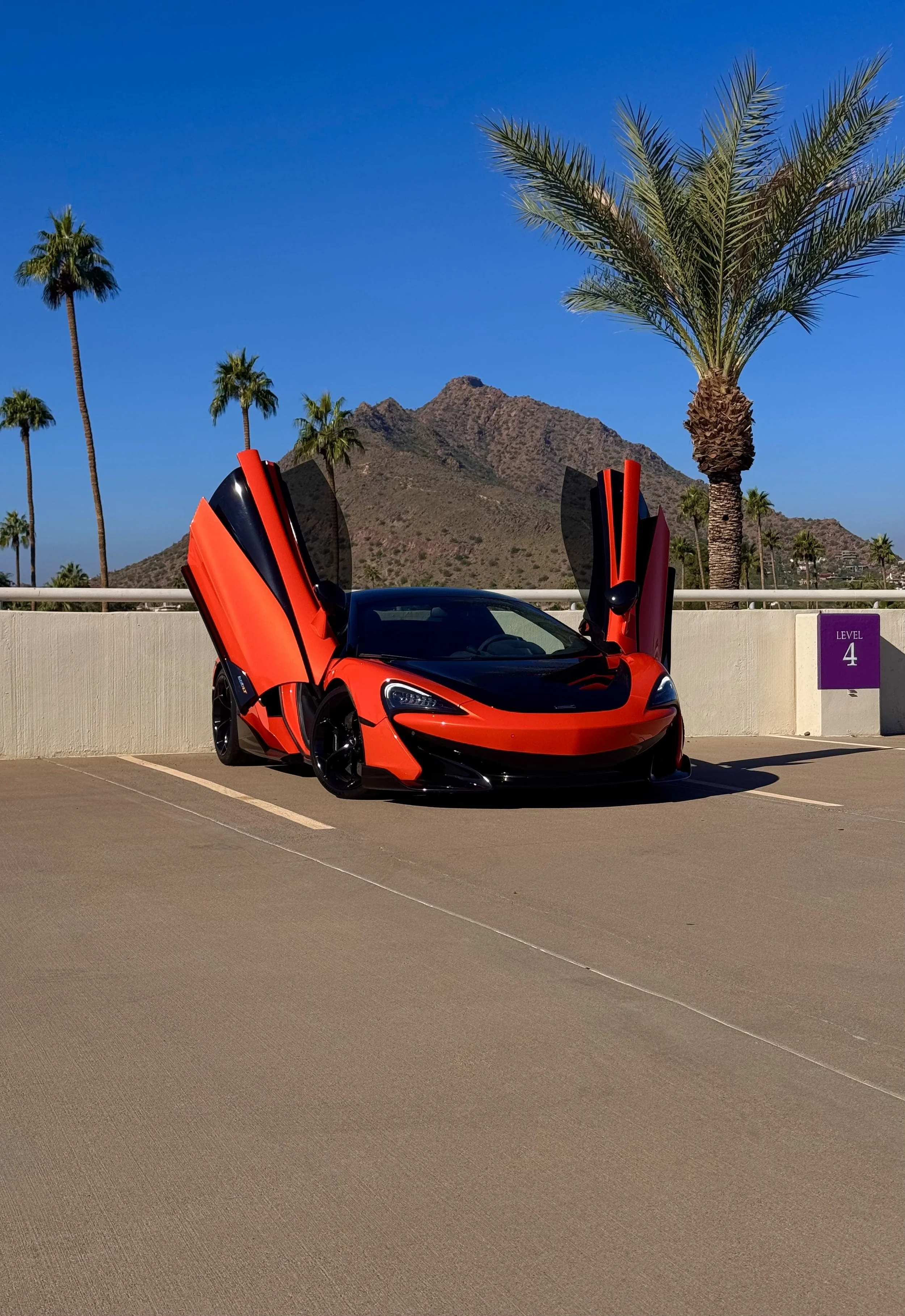 A tuned and straight-piped Helios orange McLaren 600LT spider exotic sports car with butterfly doors open, parked in an outdoor lot with palm trees and mountains in the background on a sunny day.