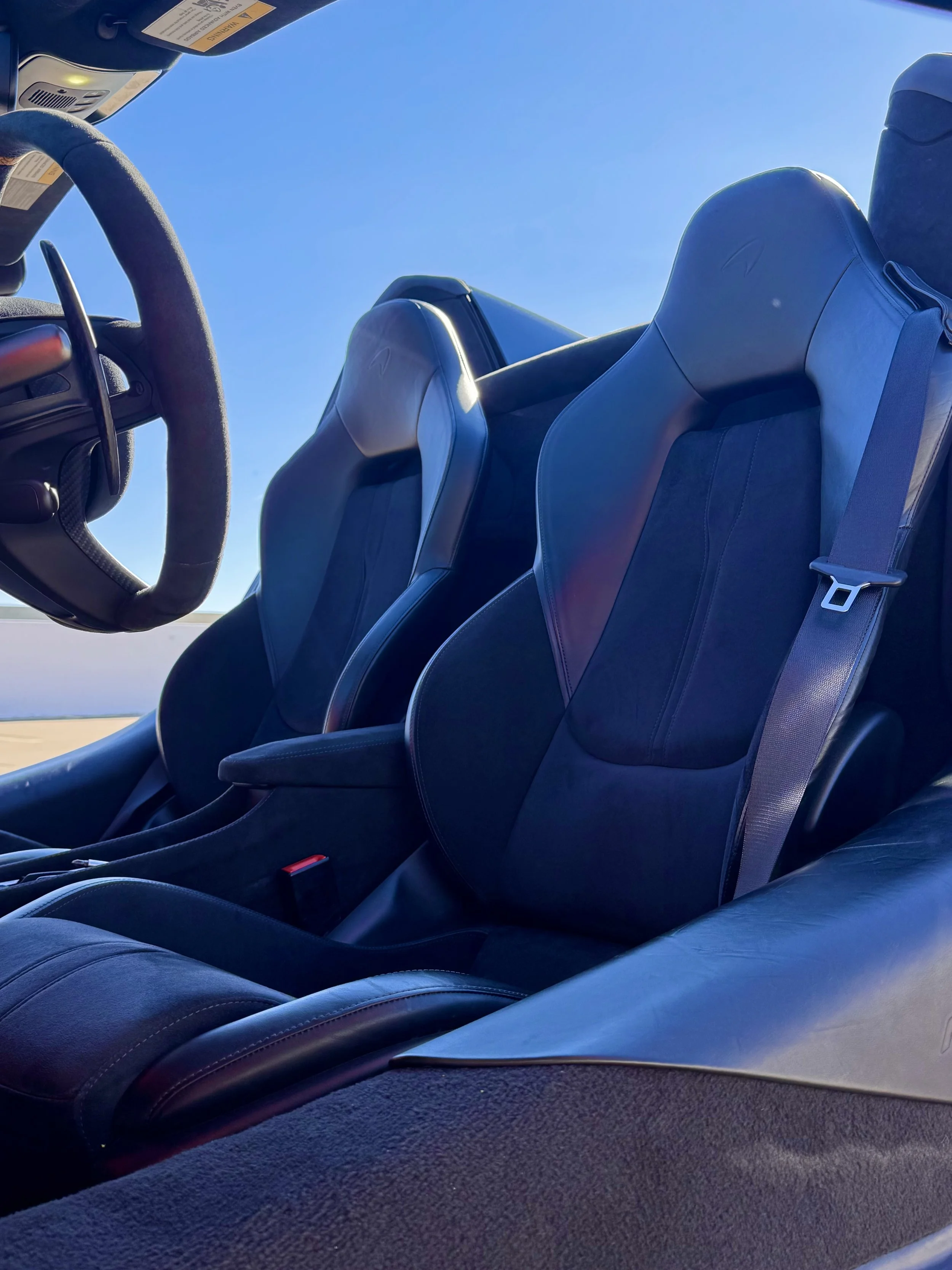 Inside the cockpit of a tuned and straight-piped Helios orange McLaren 600LT spider exotic sports car, showing black leather seats and a steering wheel, with a clear blue sky visible through the windshield.