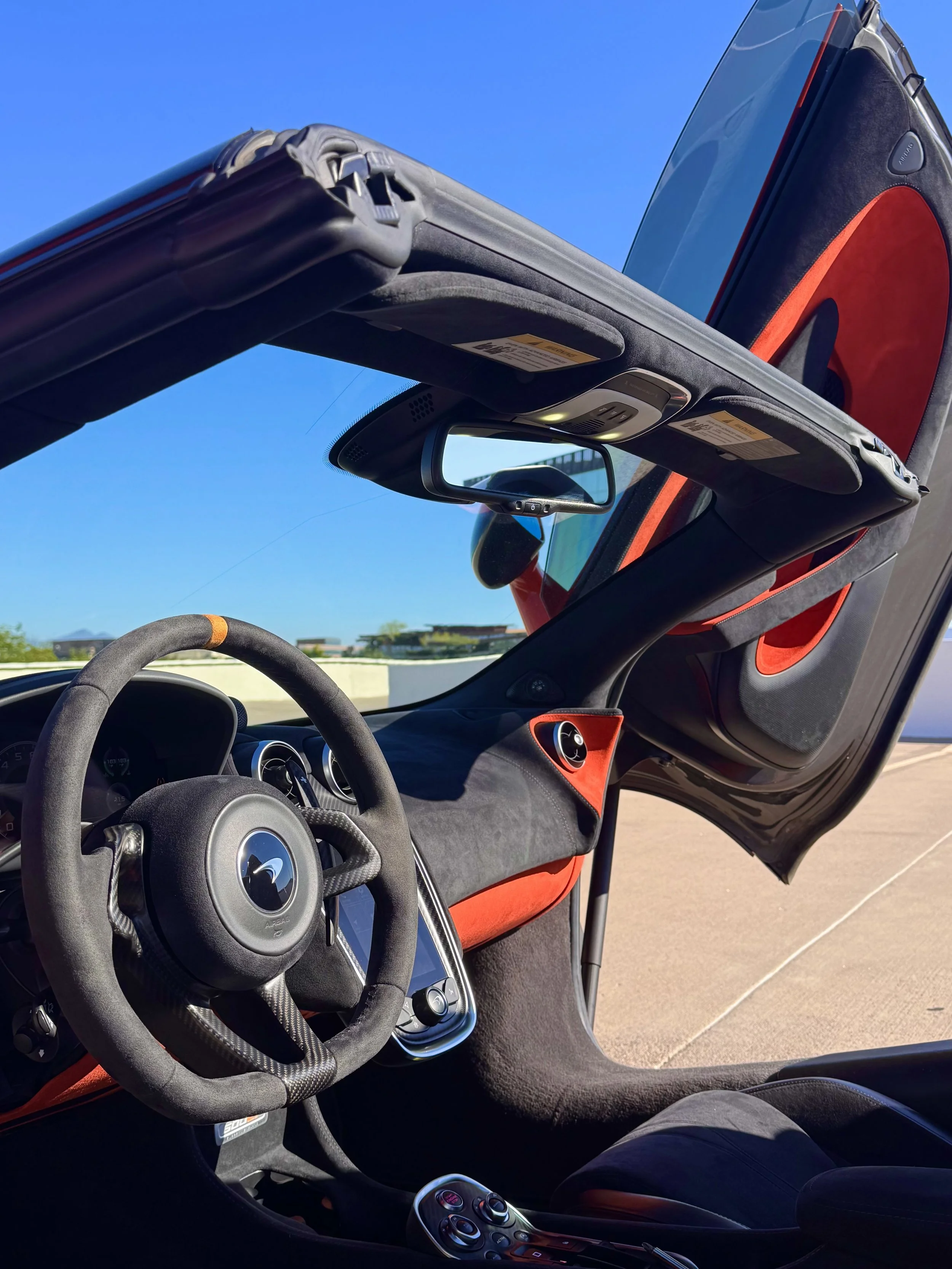 Interior of a tuned and straight-piped Helios orange McLaren 600LT spider exotic sports car with orange and black alcantara, an alcantara steering wheel with a McLaren logo, and a removable roof panel removed, showing a clear blue sky outside.