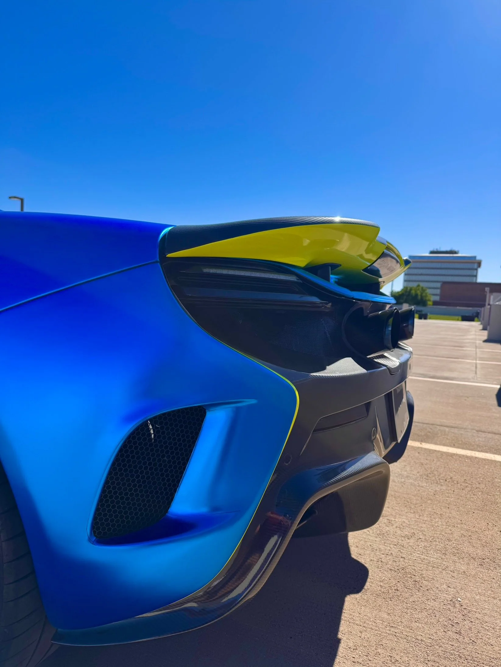 Close-up of the rear side of a blue McLaren 650S Spider with yellow accents parked outdoors on a sunny day.