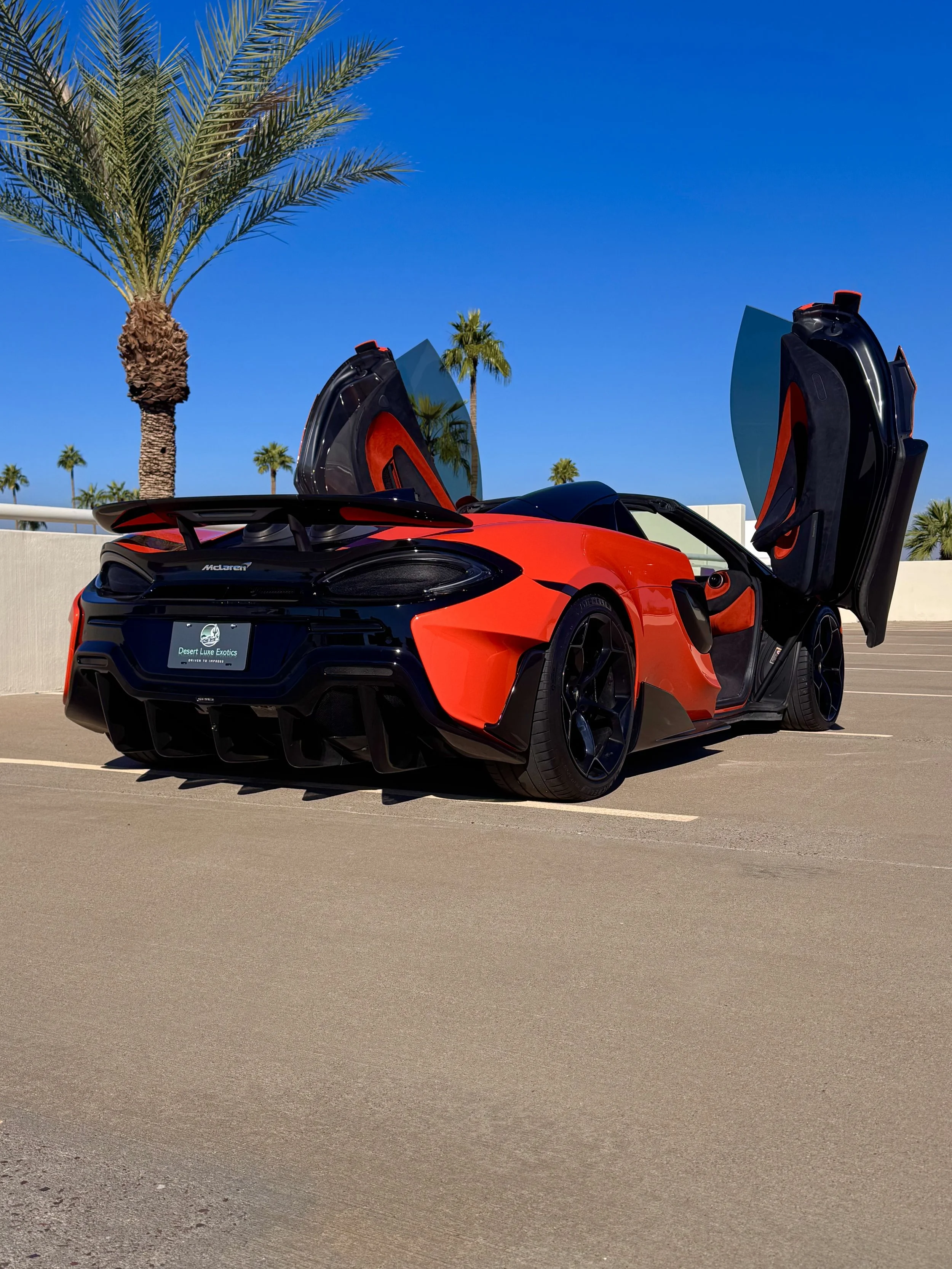 A tuned and straight-piped Helios orange McLaren 600LT spider exotic sports car with butterfly doors open parked in a sunny, outdoor lot with palm trees in the background.