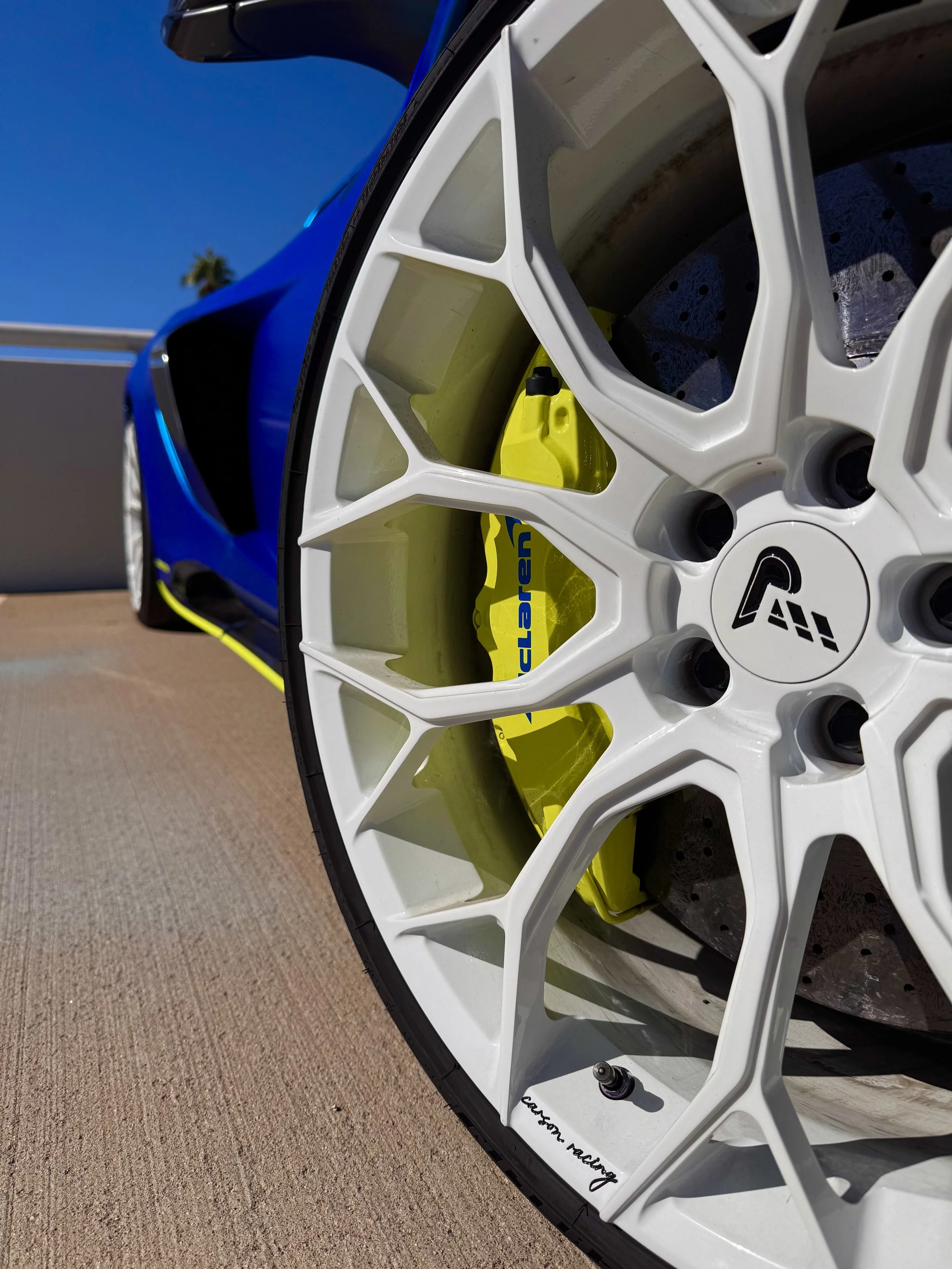 Close-up of a white wheel rim of a blue McLaren 650S Spider, revealing a yellow caliper with 'Clearspeed' written on it and a black logo center cap, on a pavement surface with a sunny clear sky in the background.