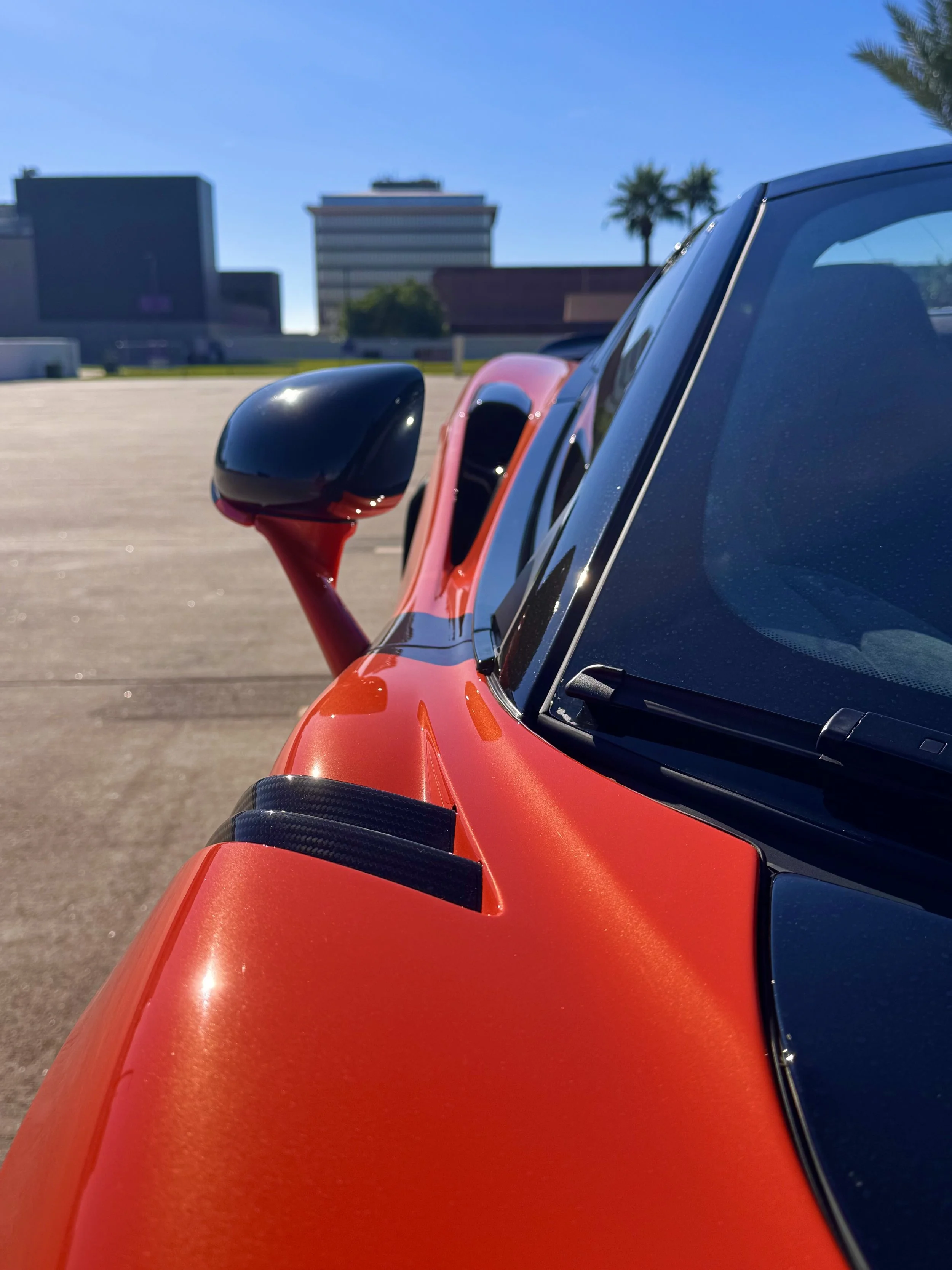 Close-up of the front part of a tuned and straight-piped Helios orange McLaren 600LT spider exotic sports car, showing the side mirror, windshield, and part of the hood, with a blurred background of buildings and palm trees under a clear blue sky.