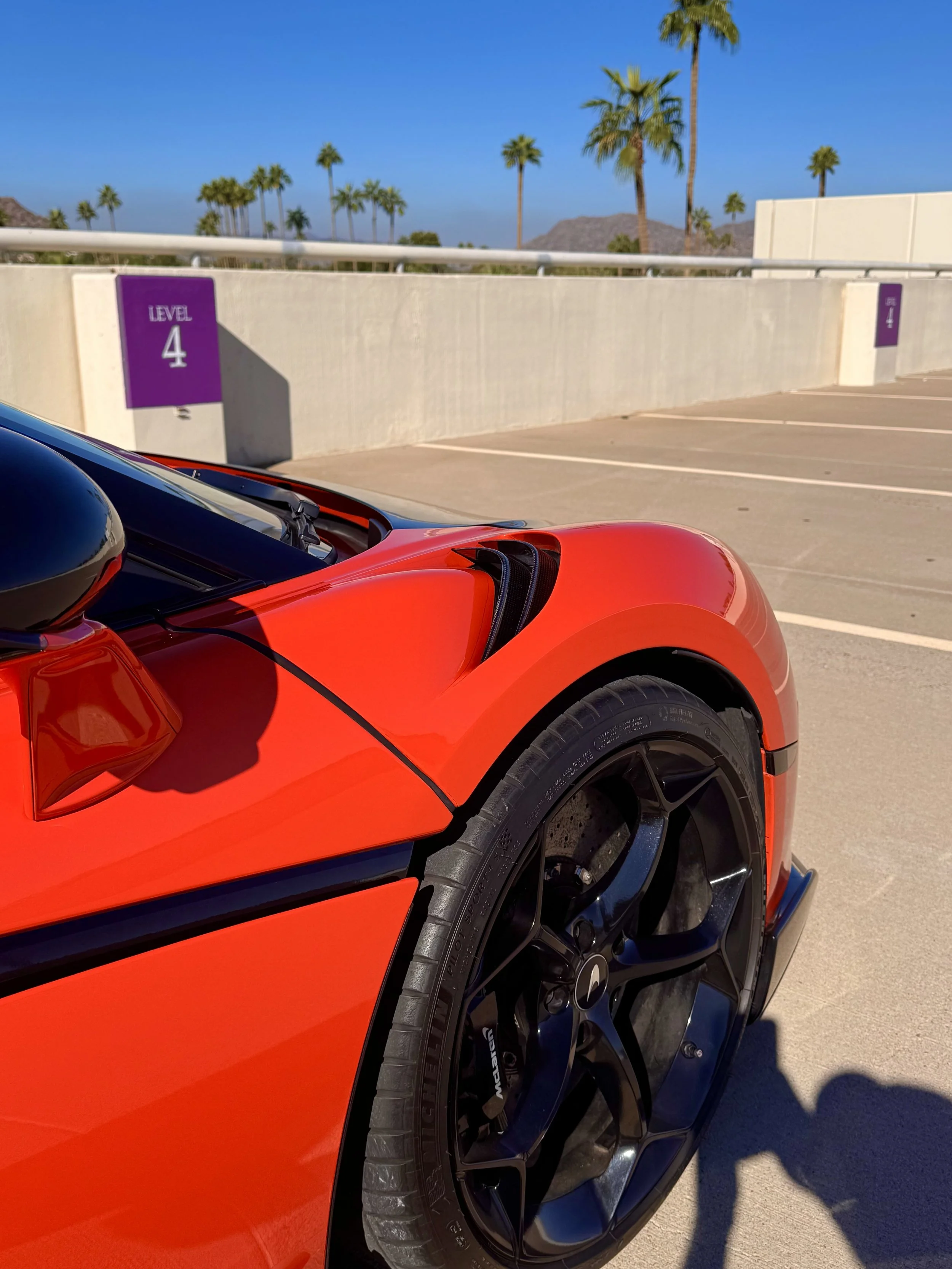 A close-up of the front part of a tuned and straight-piped Helios orange McLaren 600LT spider exotic sports car parked in a multi-story parking garage with a purple 'Level 4' sign in the background.