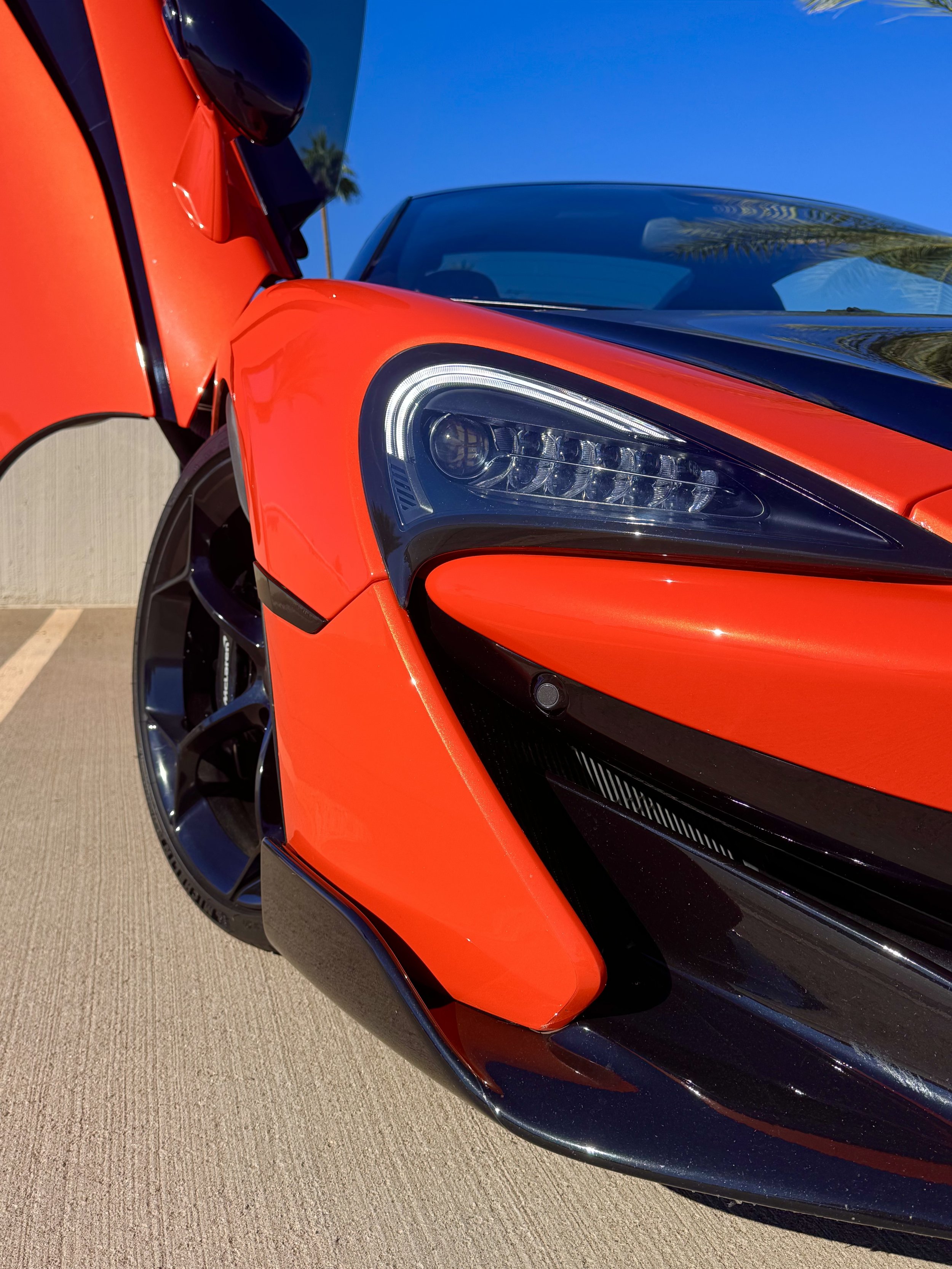 Close-up of a tuned and straight-piped Helios orange McLaren 600LT spider exotic sports car with its door open, showing the headlight and front wheel against a bright blue sky with palm trees in the background.