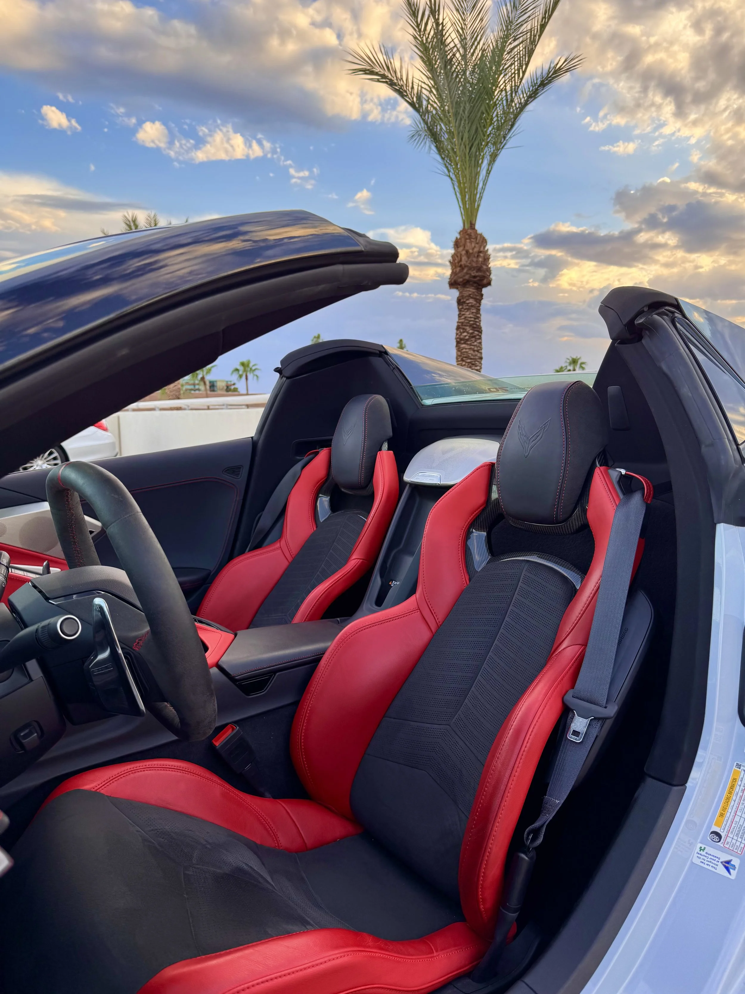 Interior of a White Chevrolet Corvette C8 convertible sports car with red and black seats, palm trees outside, and a partly cloudy sky.