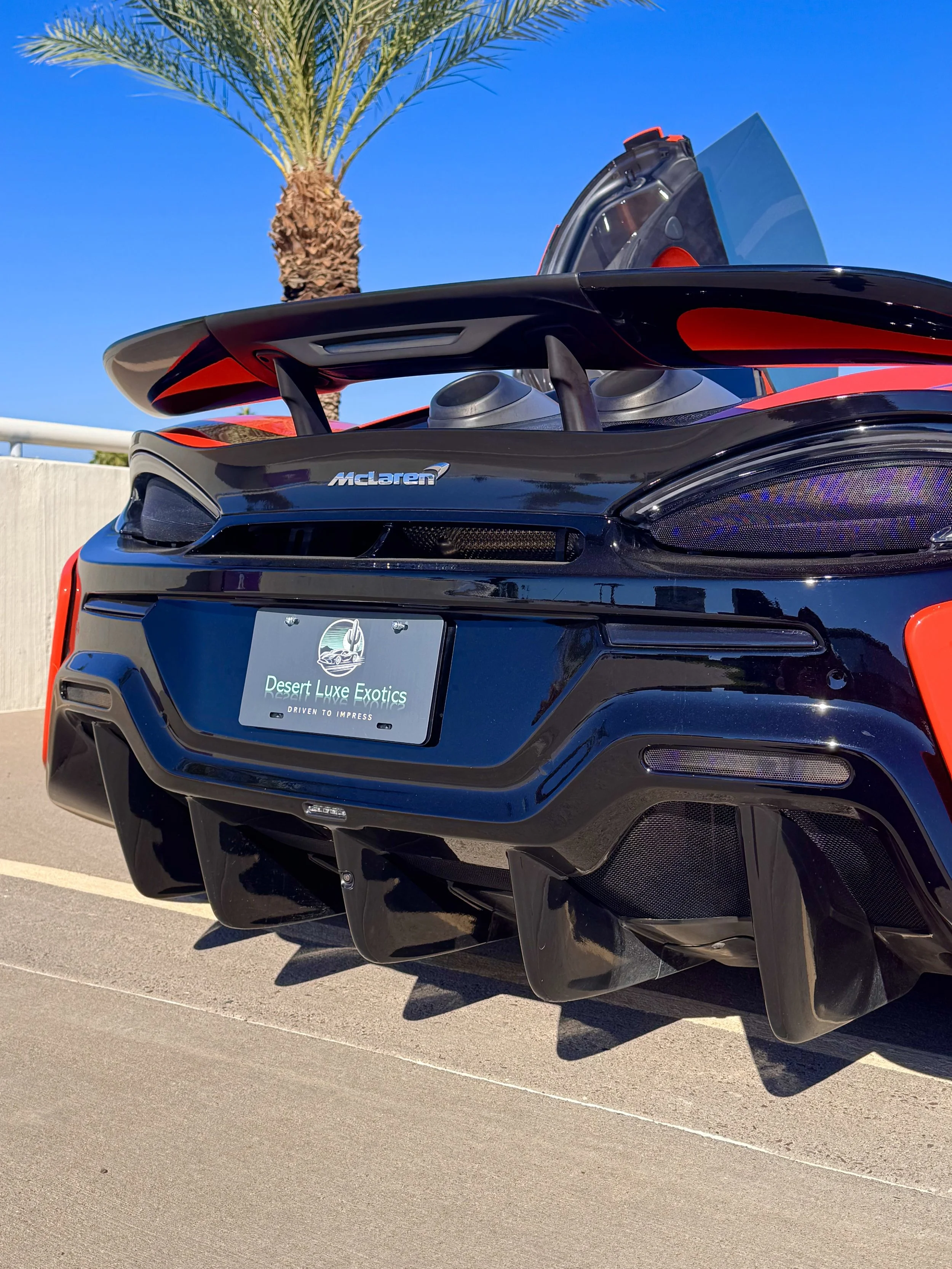 Rear view of a tuned and straight-piped Helios orange McLaren 600LT spider exotic sports car with an orange alcantara interior, license plate reading 'Desert Luxe Exotics,' parked on a sunny day with a palm tree and clear blue sky in the background.