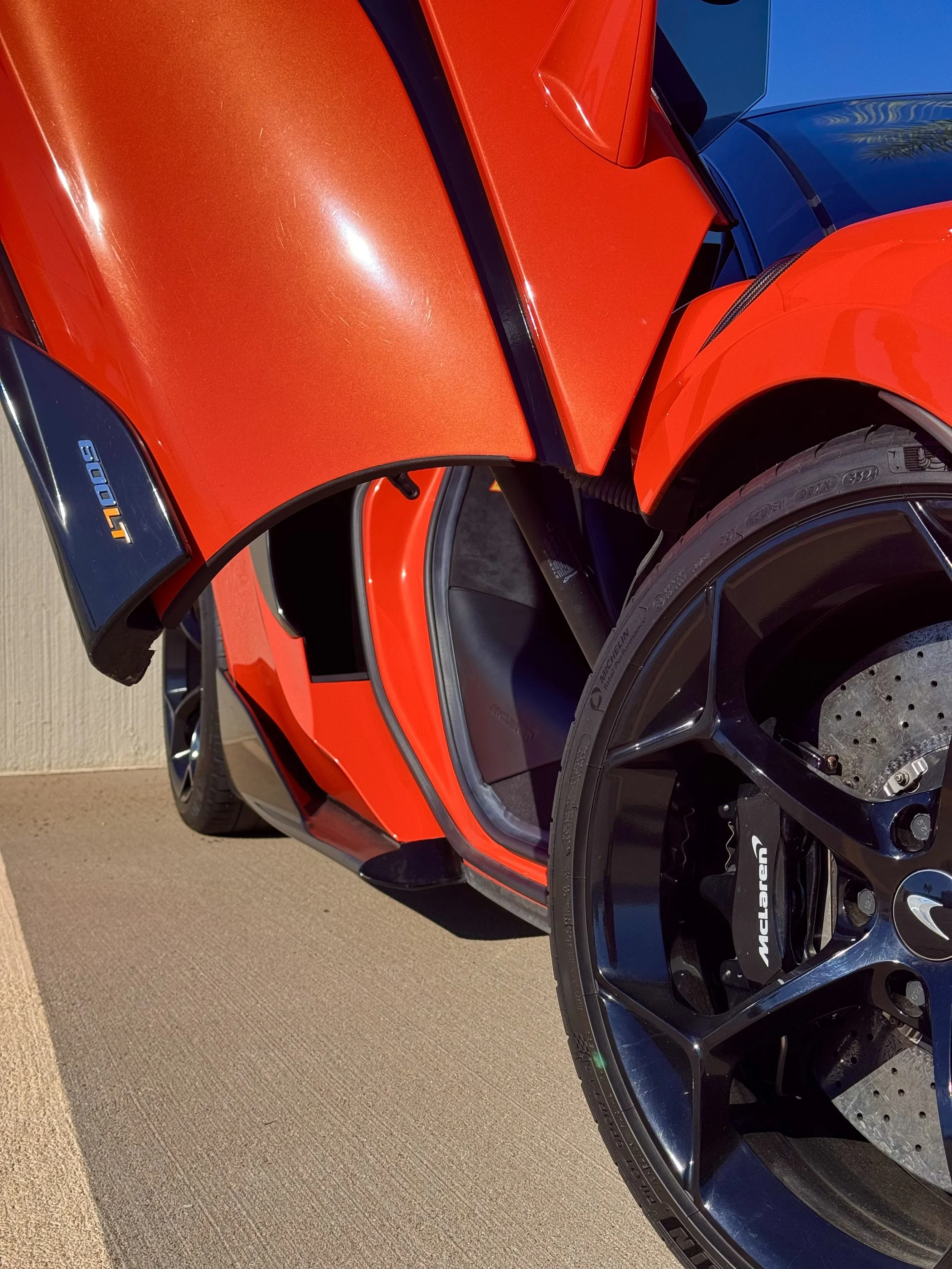 Close-up of a tuned and straight-piped Helios orange McLaren 600LT spider exotic sports car with its butterfly door open, showing the interior and the front wheel.