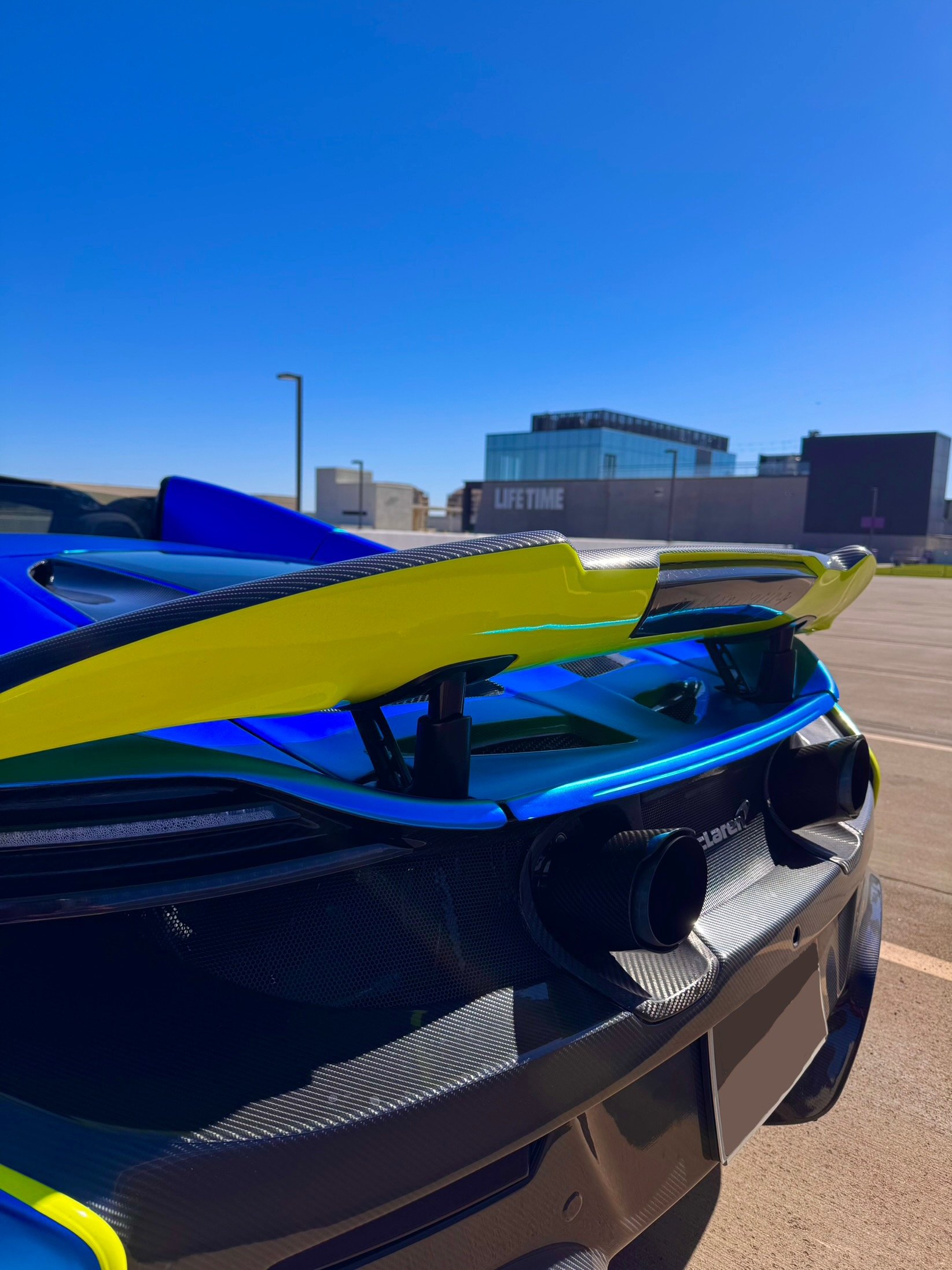 Close-up of a blue McLaren 650S Spider's rear, showing a large rear wing, dual exhaust pipes, and the McLaren logo, parked in an outdoor lot with a modern building and clear blue sky in the background.
