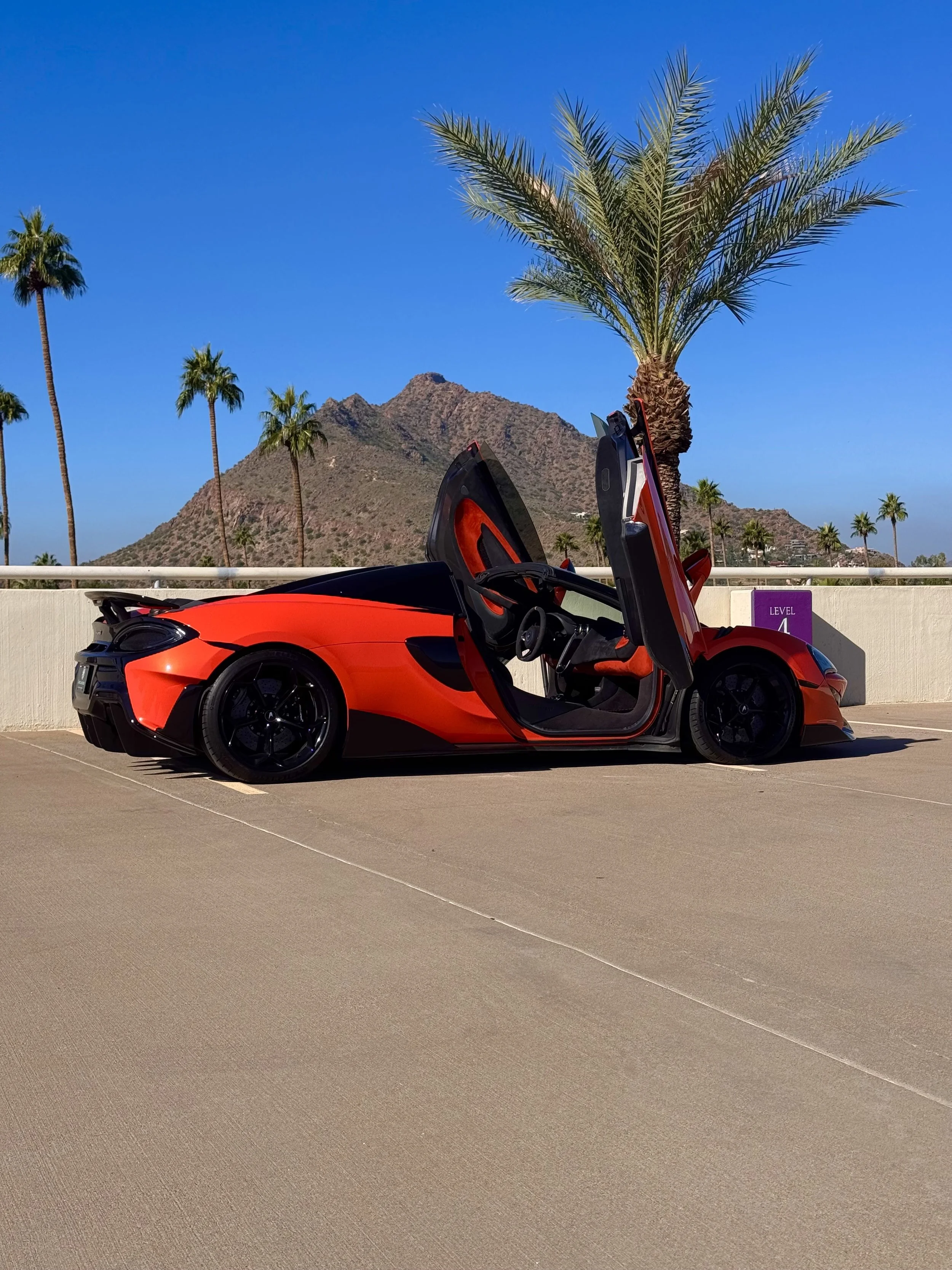 A tuned and straight-piped Helios orange McLaren 600LT spider exotic sports car with butterfly doors open, parked on a rooftop with palm trees and mountains in the background.
