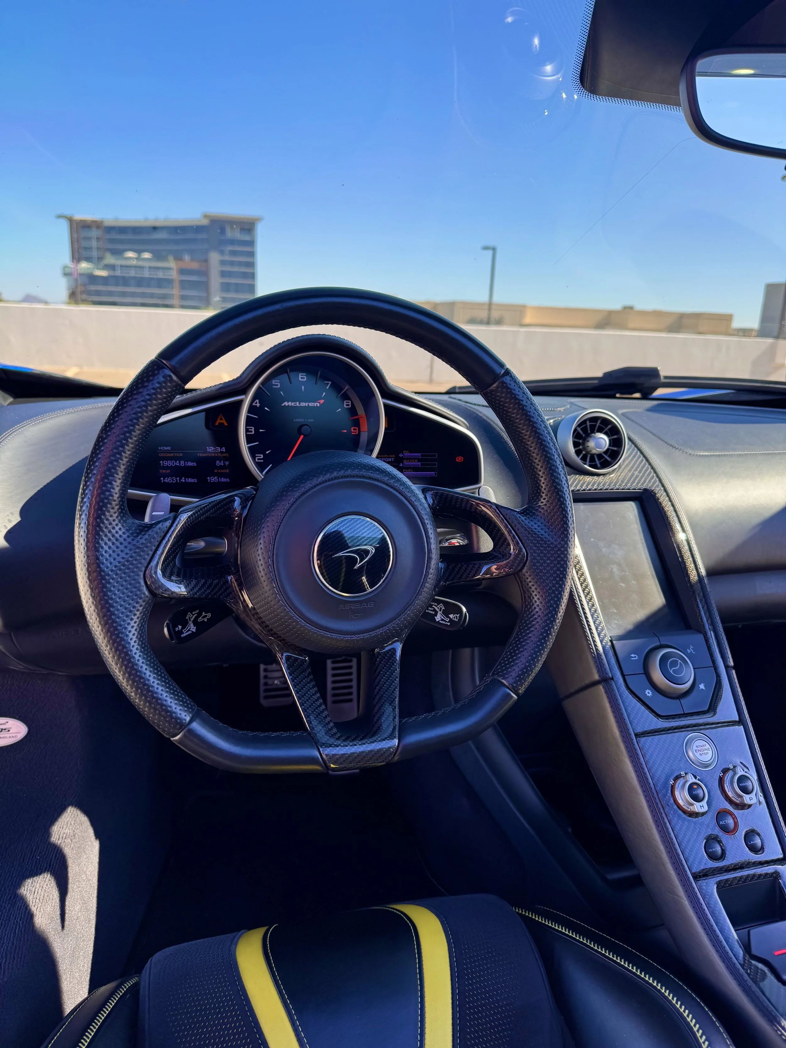 Interior view of a blue McLaren 650S Spider showing the steering wheel, instrument cluster, and center console with controls, with buildings and a clear blue sky visible through the windshield.