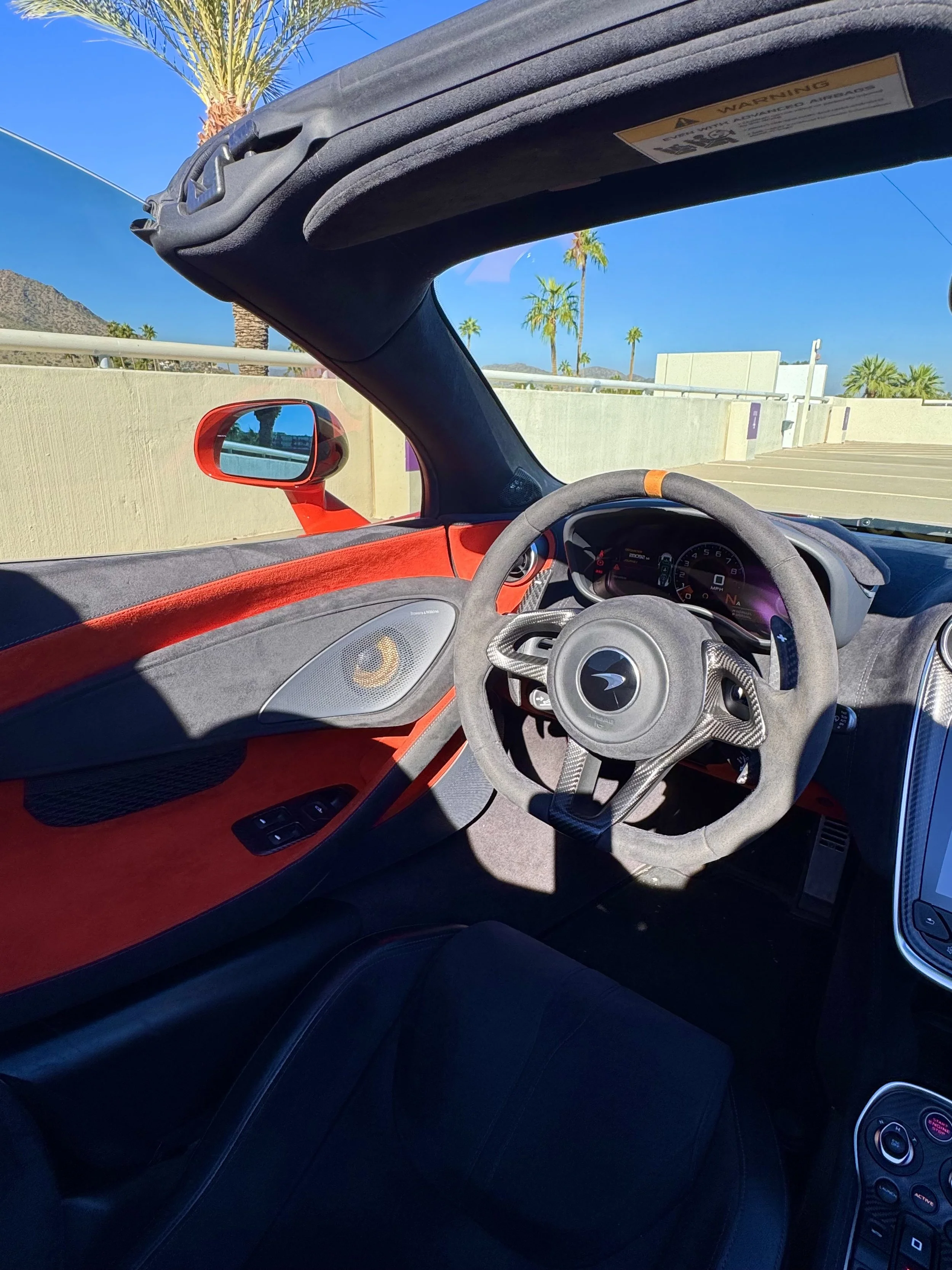 Interior of a tuned and straight-piped Helios orange McLaren 600LT spider exotic sports car with a carbon fiber steering wheel, showing the dashboard and side mirror, with palm trees and a blue sky outside.