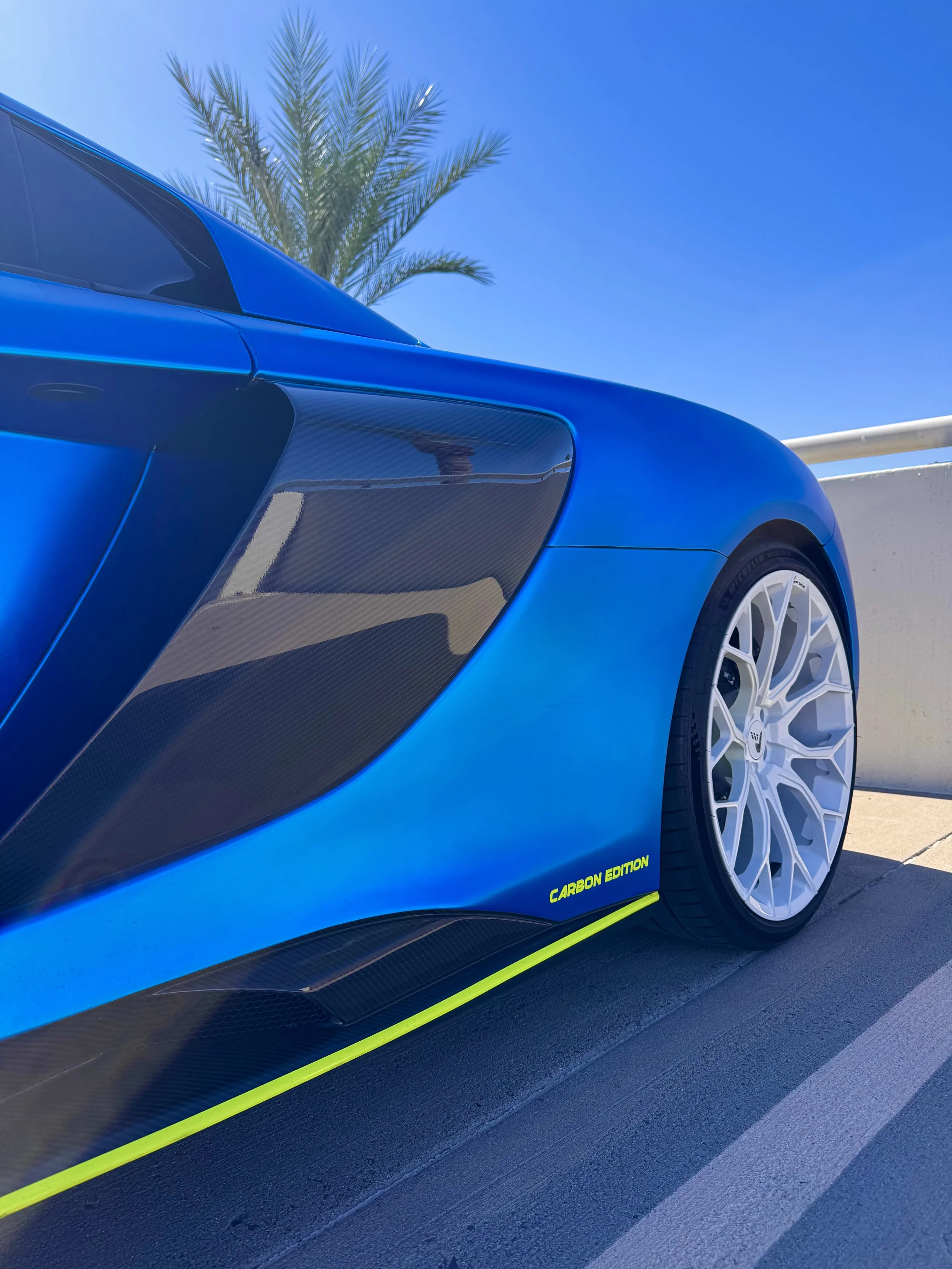 Close-up of a blue McLaren 650S Spider with carbon fiber accents, a white alloy wheel, and a yellow stripe with the text 'Carbon Edition', parked outdoors under a clear blue sky with a palm tree in the background.
