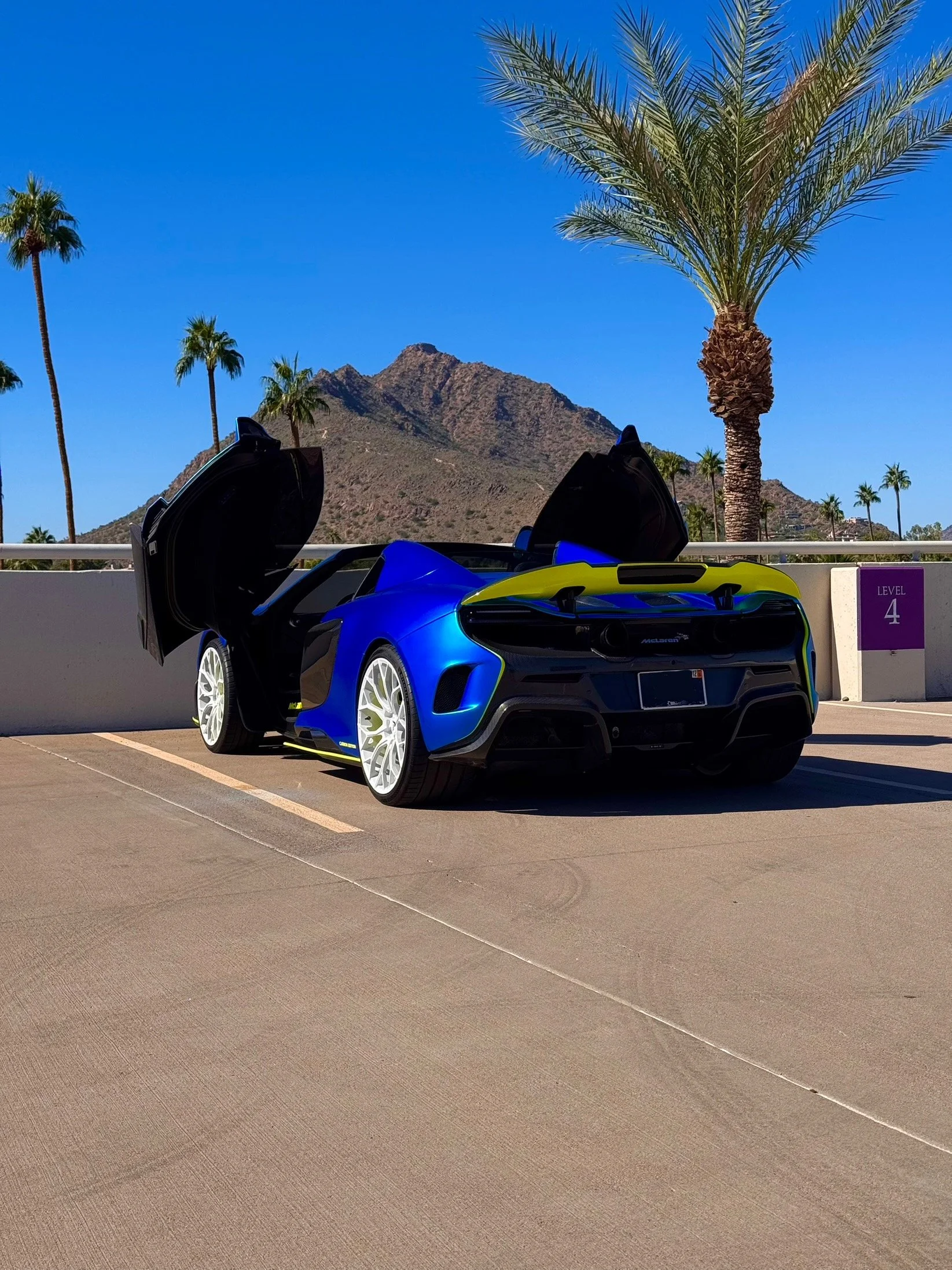 A blue McLaren 650S Spider with butterfly doors open parked in a sunny outdoor parking lot with palm trees, mountains, and a clear blue sky in the background.