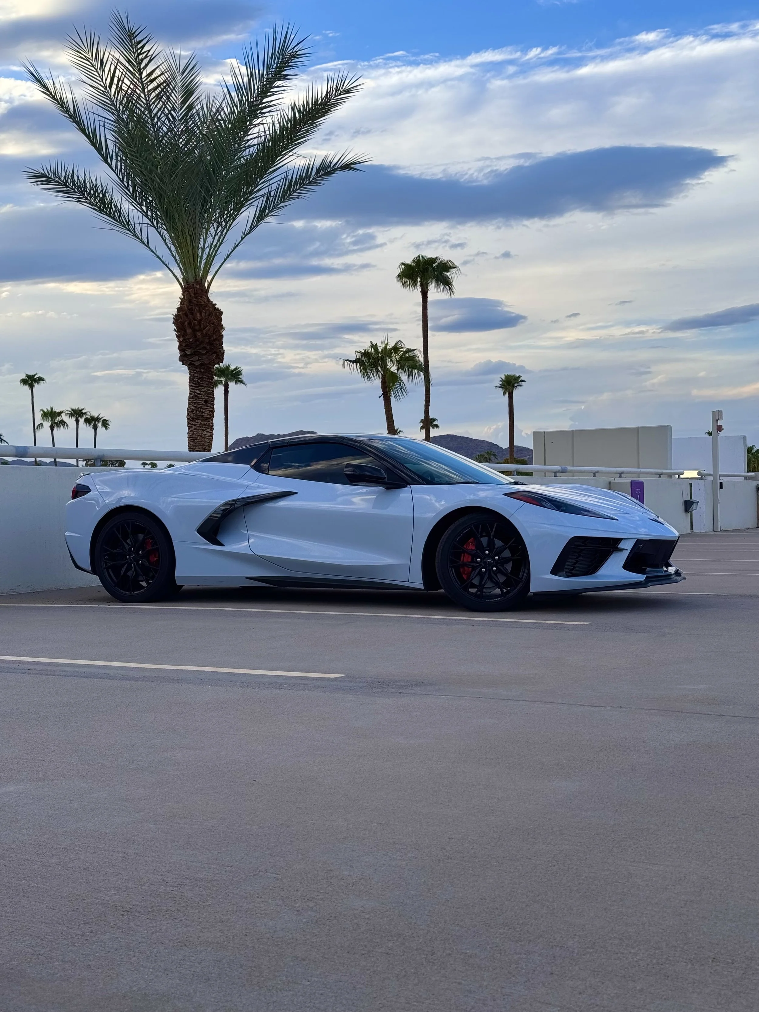 A White Chevrolet Corvette C8 convertible sports car parked on a concrete road with palm trees and mountains in the background under a partly cloudy sky.