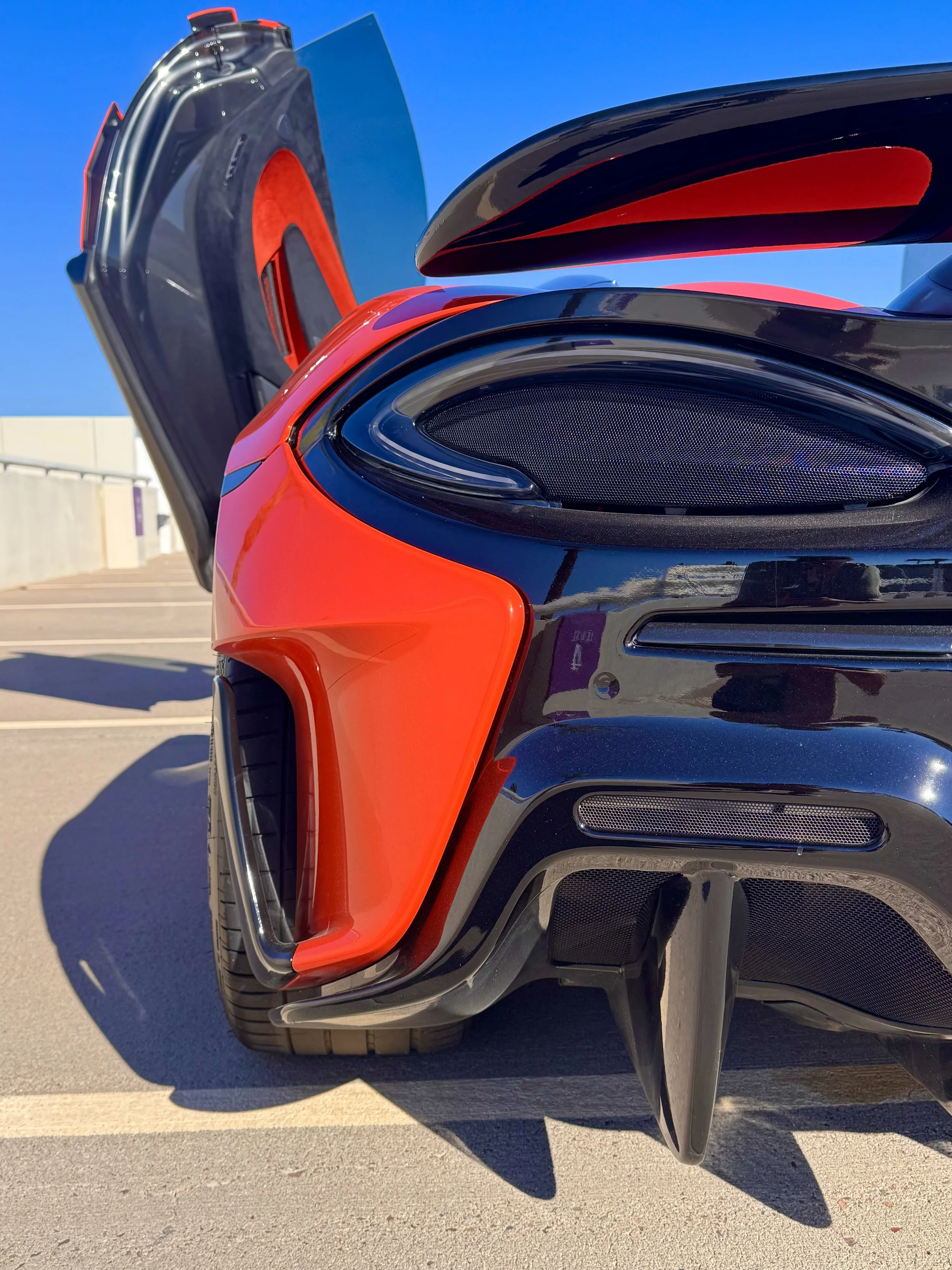 Close-up of the rear end of a tuned and straight-piped Helios orange McLaren 600LT spider exotic sports car with its rear wing open, parked on a rooftop under a clear blue sky.
